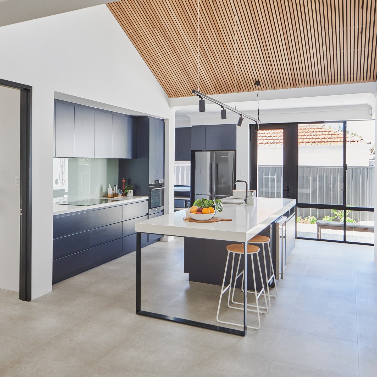Open-plan modern kitchen featuring matte navy blue cabinets, a white island with a steel frame, and a dramatic pitched ceiling clad in timber slats.