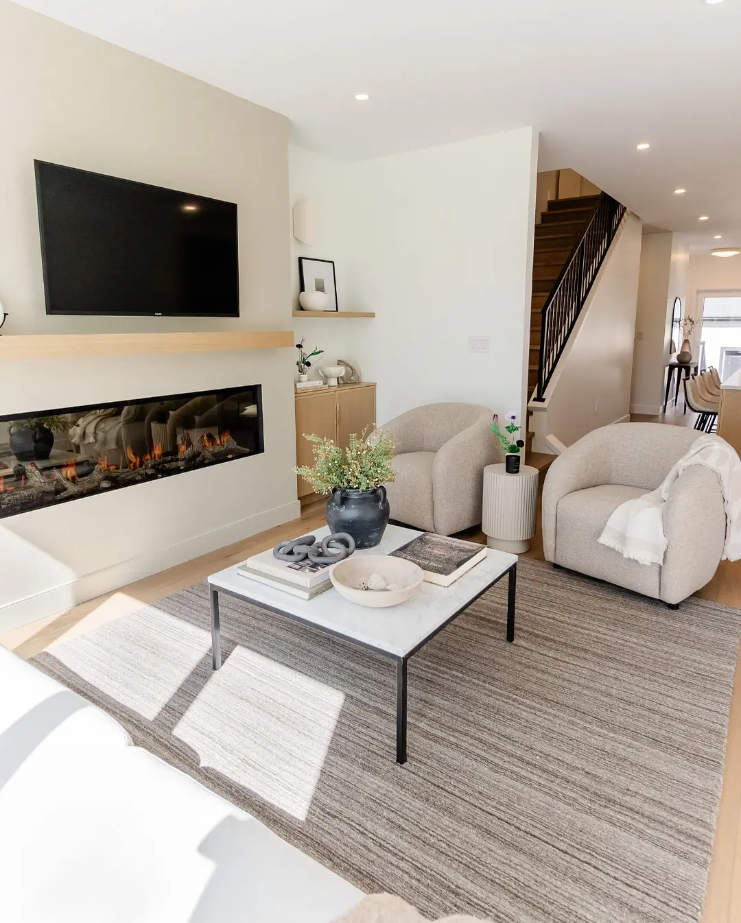 Contemporary living room with a cream-colored feature wall, a wide linear electric fireplace, a light wood floating mantel, a mounted TV, and matching light wood furniture.