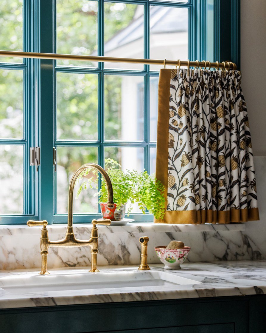 Kitchen sink with a gold faucet and marble counter, featuring a teal window frame and a cafe curtain with a black and gold botanical print and mustard yellow velvet trim.