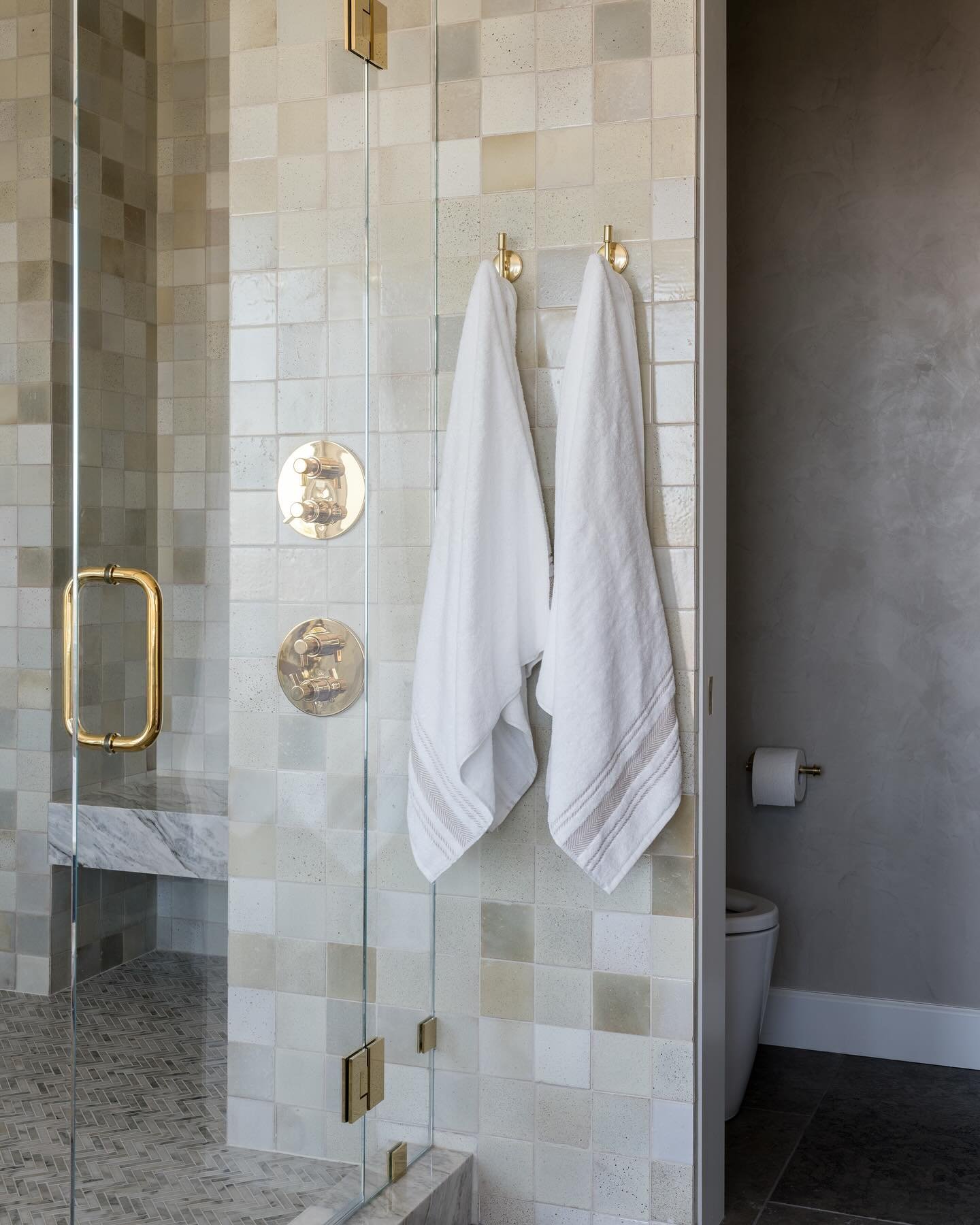 Two round brass wall hooks holding white towels, mounted on a clear glass shower partition next to brass shower controls in a tiled bathroom.