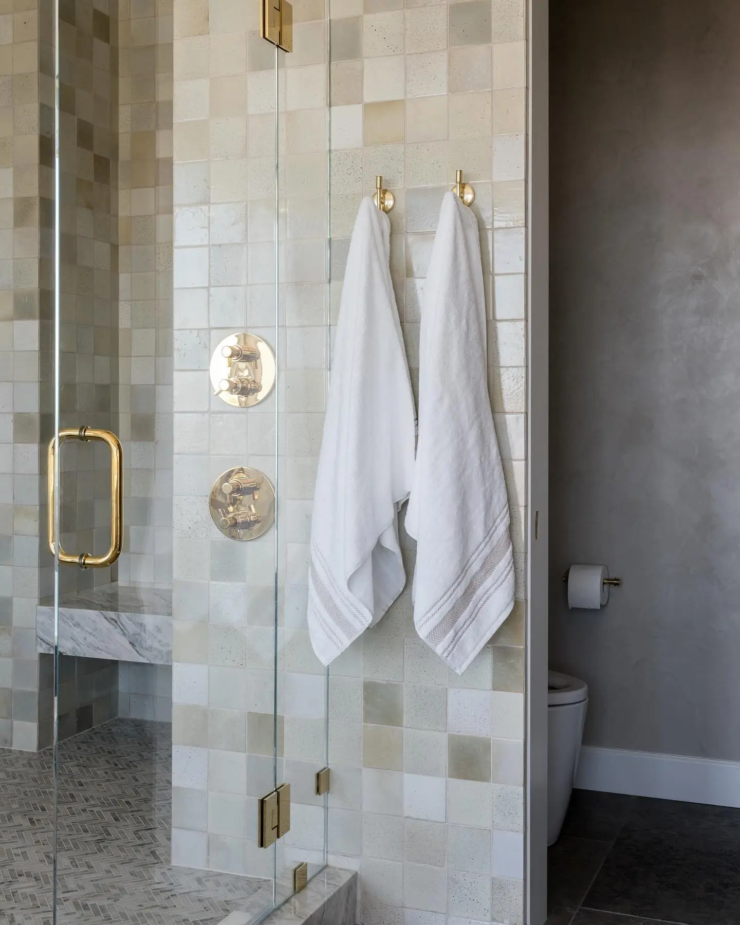 Two round brass wall hooks holding white towels, mounted on a clear glass shower partition next to brass shower controls in a tiled bathroom.