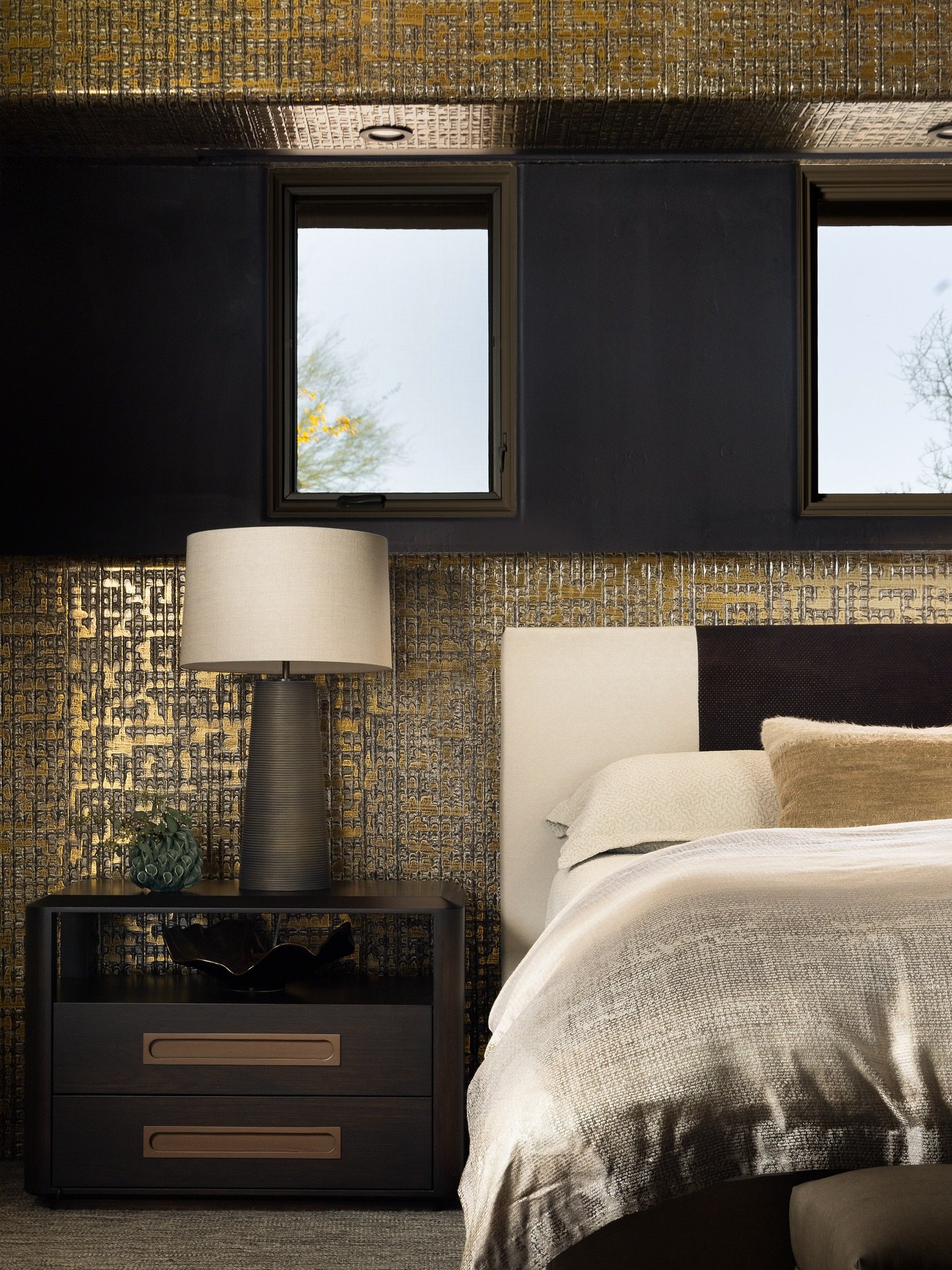 Dark, two-drawer nightstand with rectangular bronze drawer pulls next to a bed, against a highly textured gold and black wall.