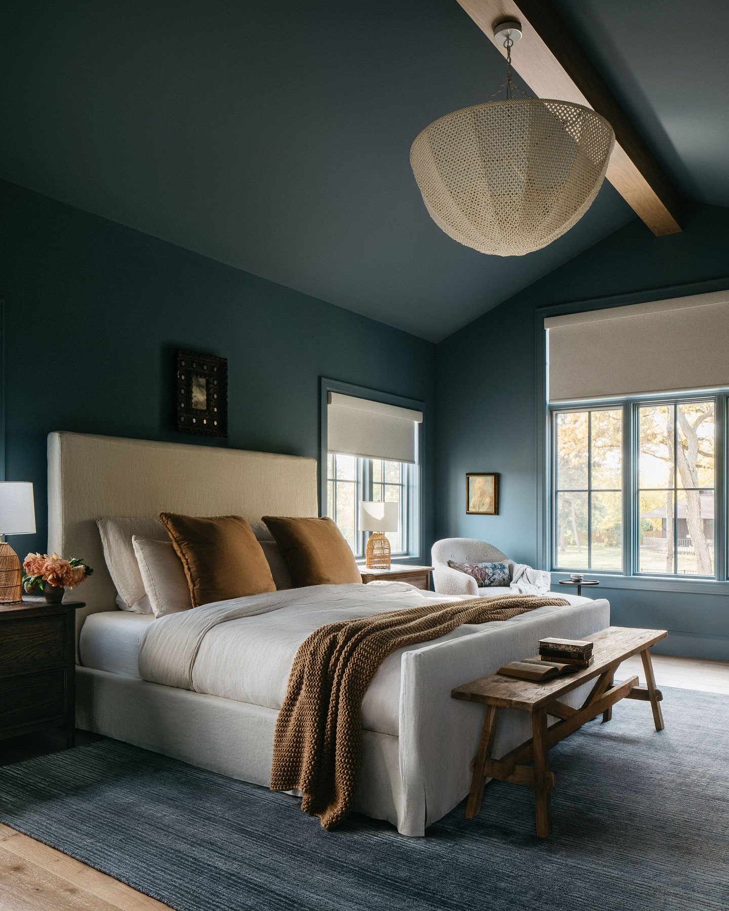 Dark bedroom painted in deep teal, featuring a vaulted ceiling with a wood beam, a large fabric upholstered bed, and terracotta accent pillows and throw.