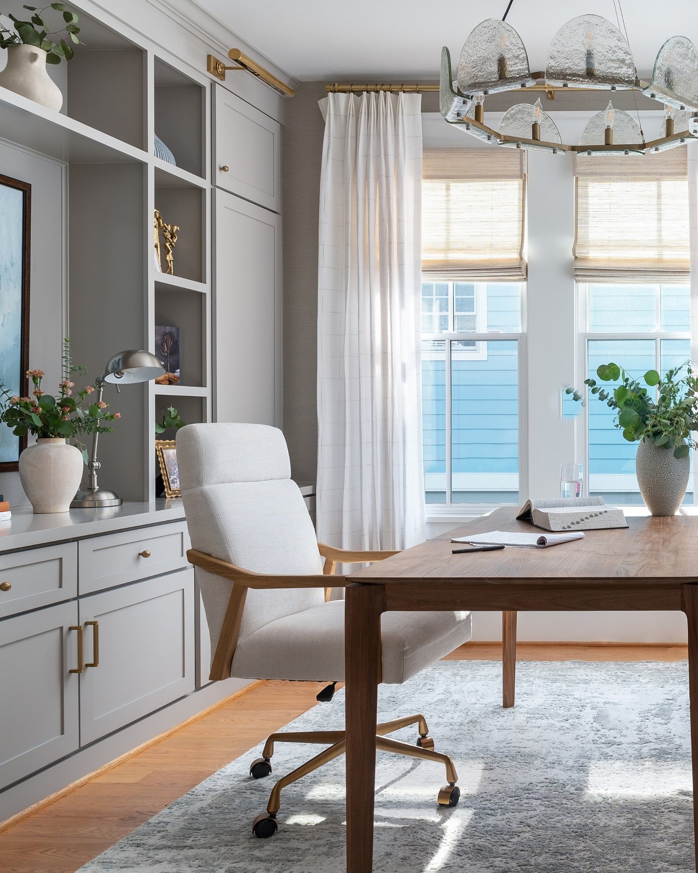 Home office with light greige built-in cabinetry, a rectangular wood desk, a white upholstered rolling chair, and a distinctive modern glass chandelier.