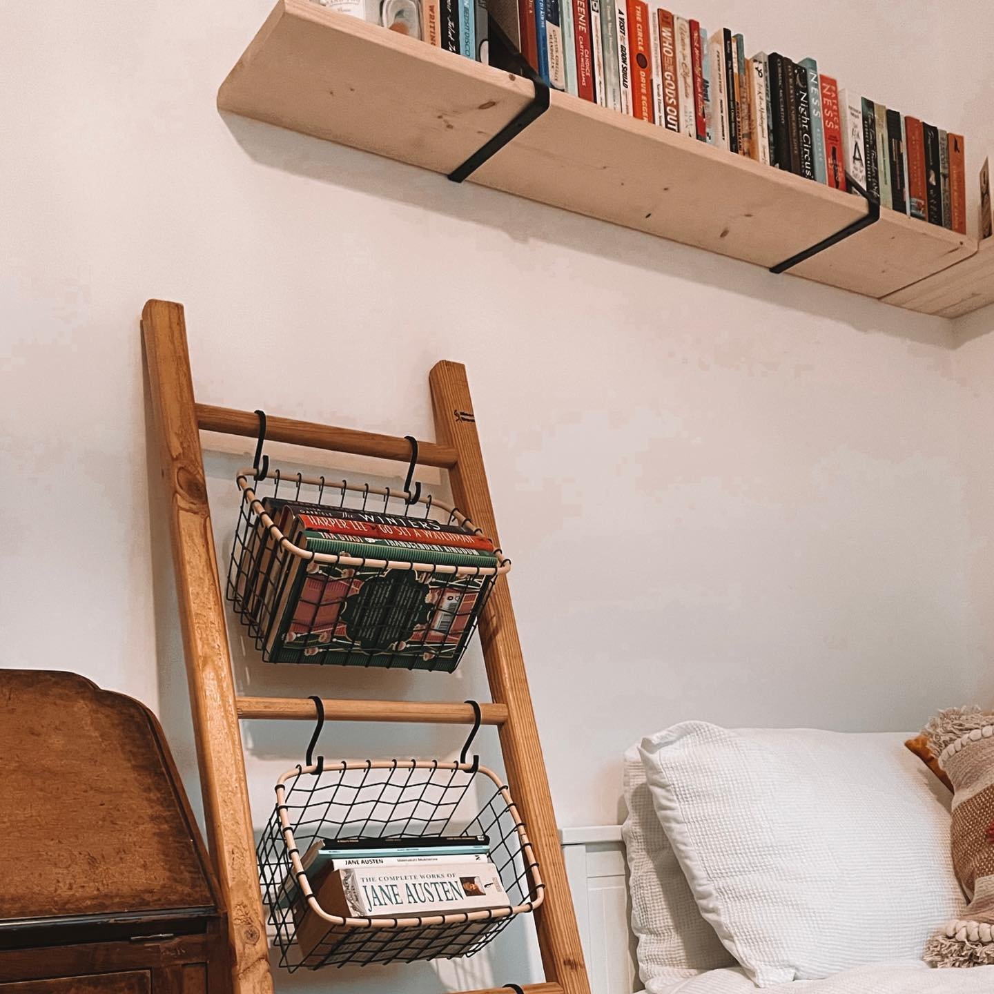 A wooden ladder leaning against a wall next to a bed, with two wire baskets hooked onto the rungs, used to store small stacks of books, with a floating natural wood shelf above.