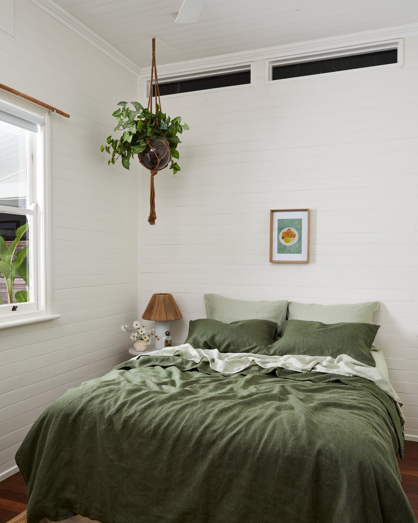 A bright bedroom with white panelled walls, a bed made with a textured olive green duvet, and a hanging plant.