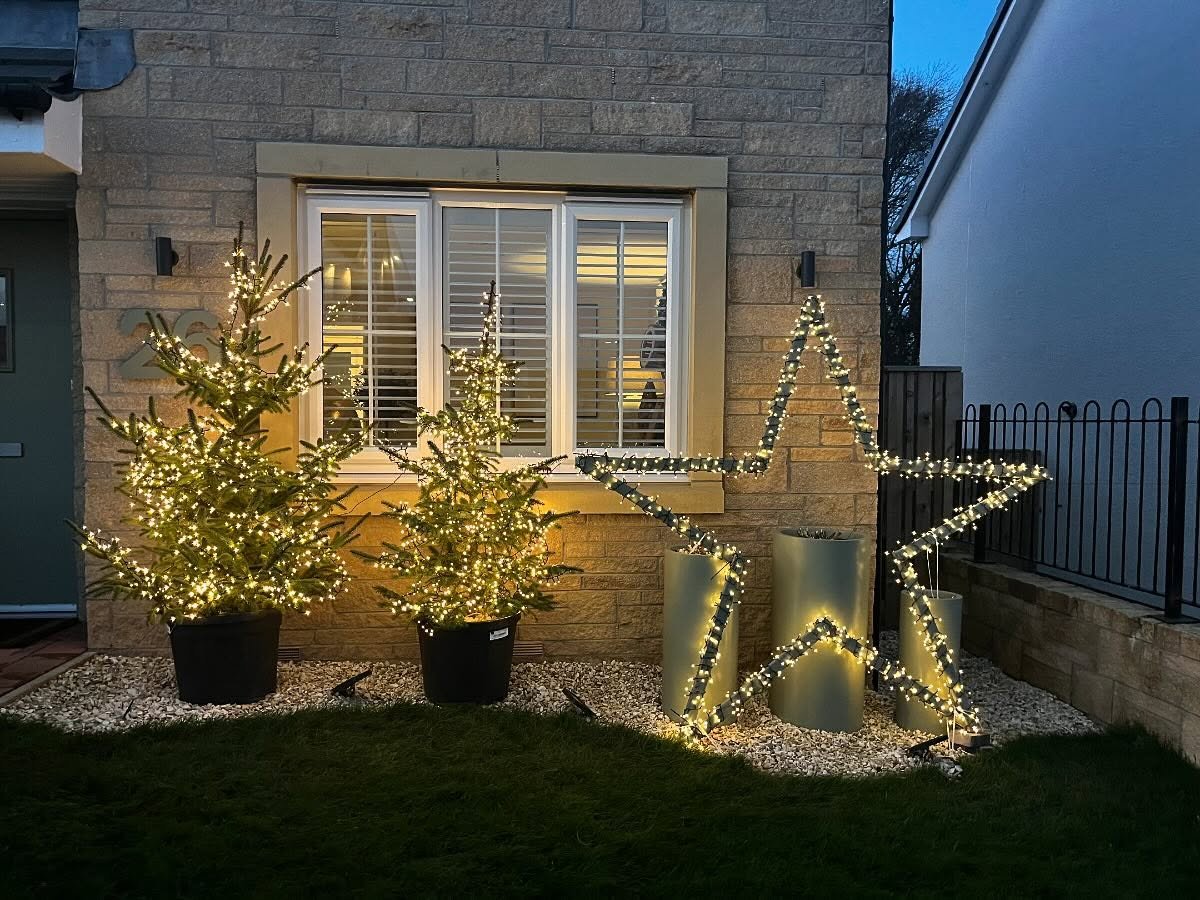 Two brightly lit potted evergreen trees sit next to a large, glowing warm white wire-frame star motif placed against a modern light stone wall beneath a window