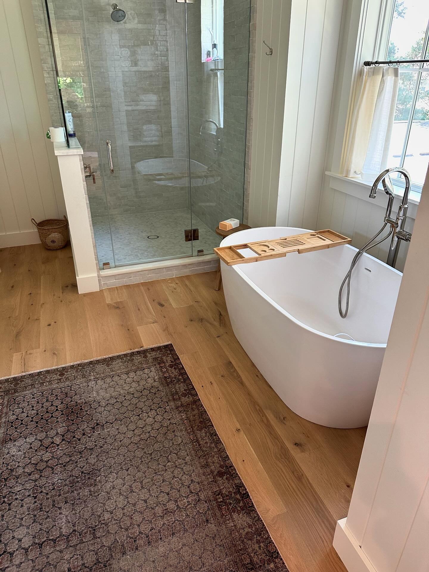 A bright bathroom featuring light wood plank flooring, white shiplap walls, a freestanding tub with a bamboo caddy, a glass walk-in shower, and a rectangular distressed dark brown and black patterned runner rug.