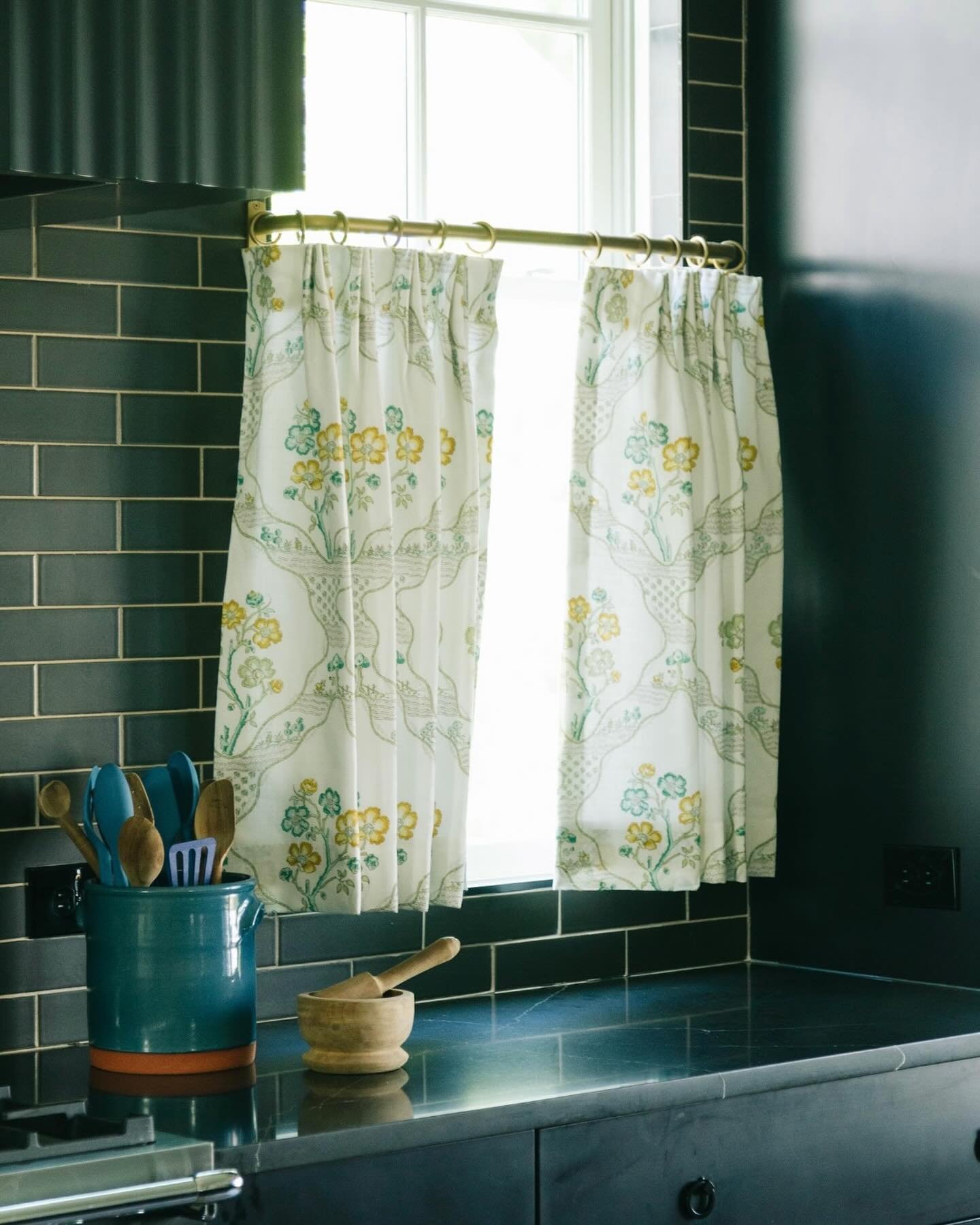 Dark kitchen with a black subway tile backsplash and counter, featuring two white cafe curtains with a yellow and teal floral pattern hung on a brass rod.