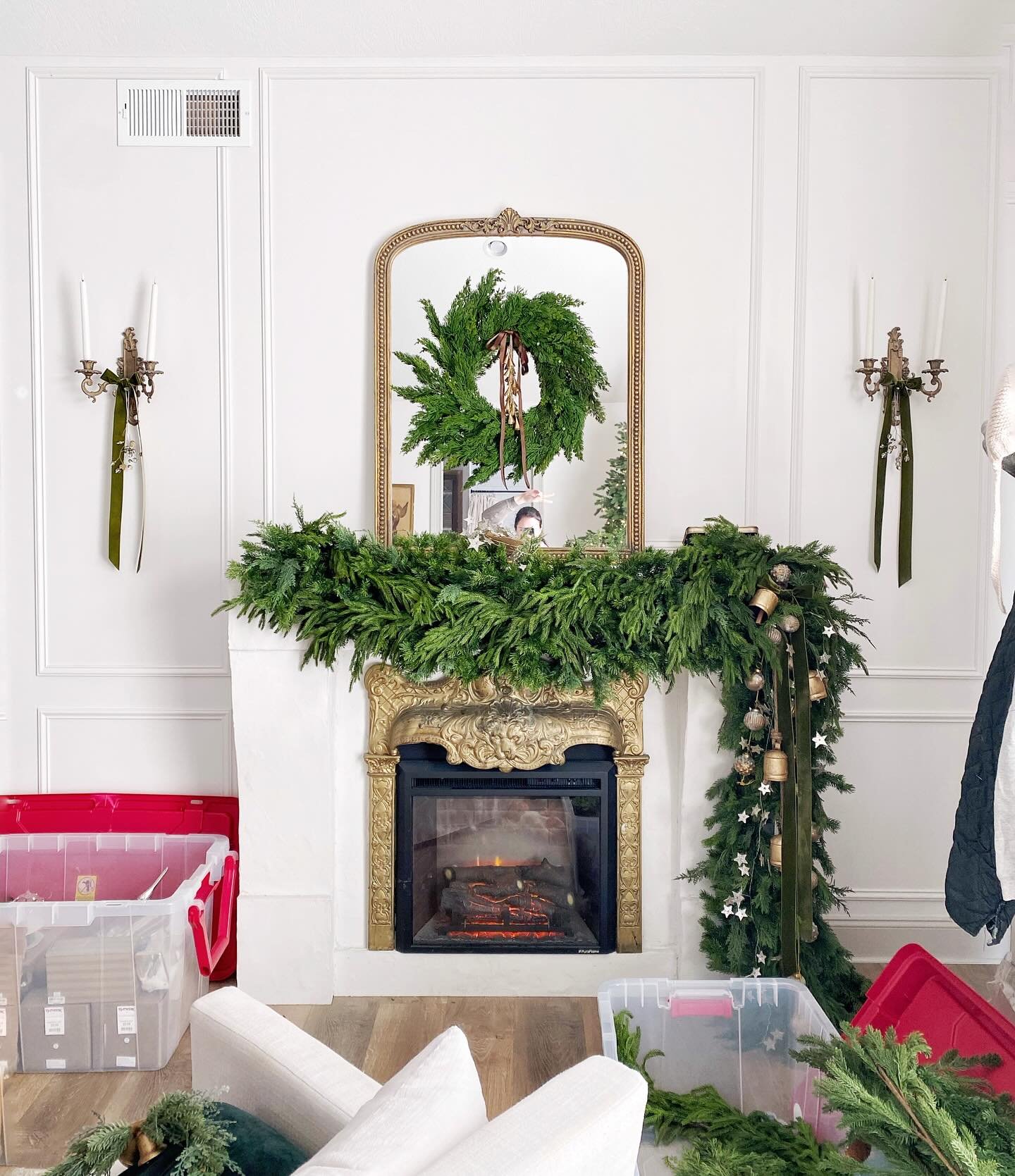 White fireplace with a gold-framed mirror, decorated with a very thick, multi-layered cedar garland, gold bells, and green velvet ribbon accents.