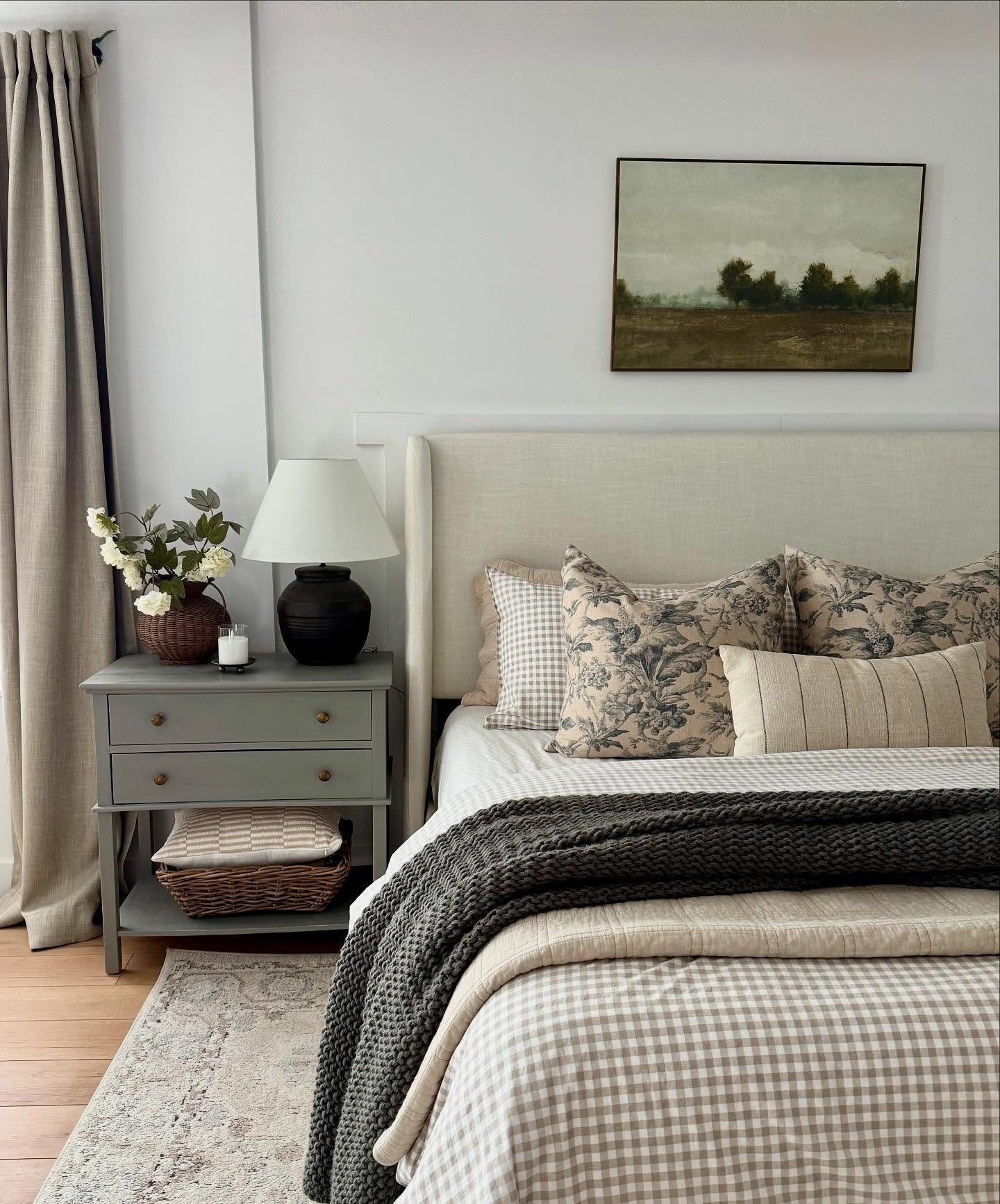 Neutral bedroom featuring a beige upholstered headboard, gingham and toile patterned bedding, a dark gray knit throw, and a gray painted nightstand.