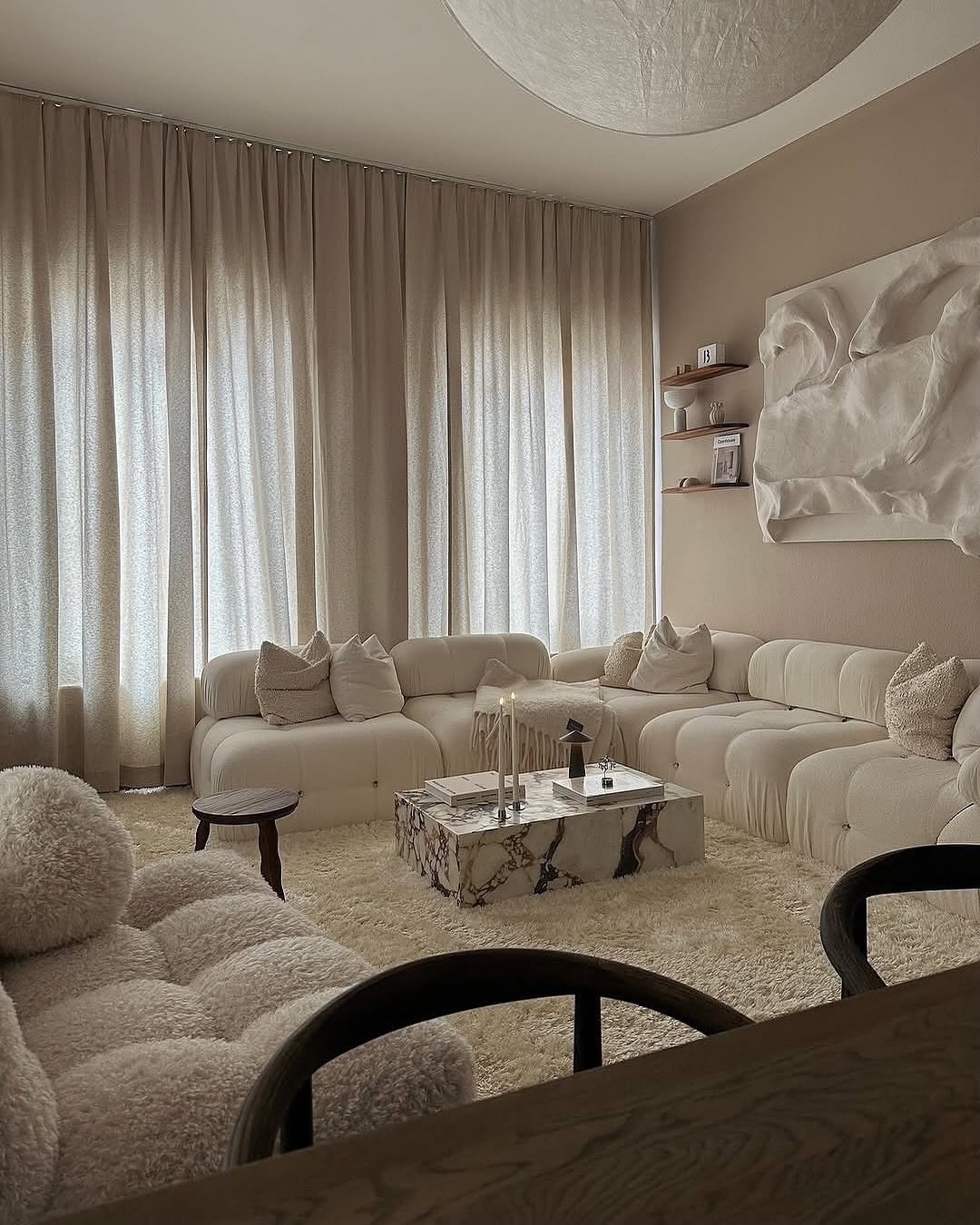 A deeply cozy, monochromatic living room with an oversized, low-slung white modular sofa, a white fluffy shag rug, sheer light beige drapery, and a marble-patterned coffee table.