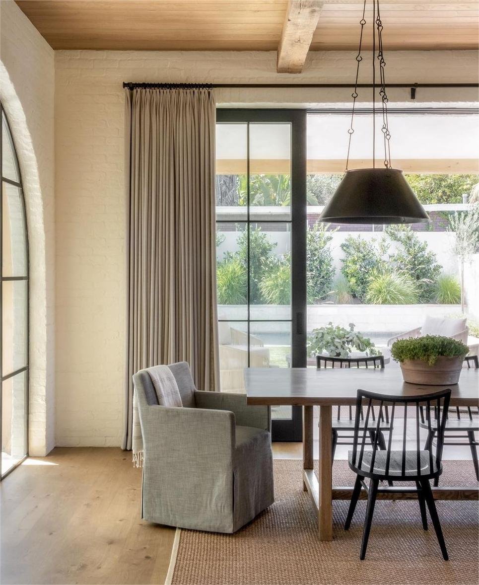 A single, heavy, taupe-colored curtain panel stacked to one side of black-framed sliding glass doors in a rustic dining area.