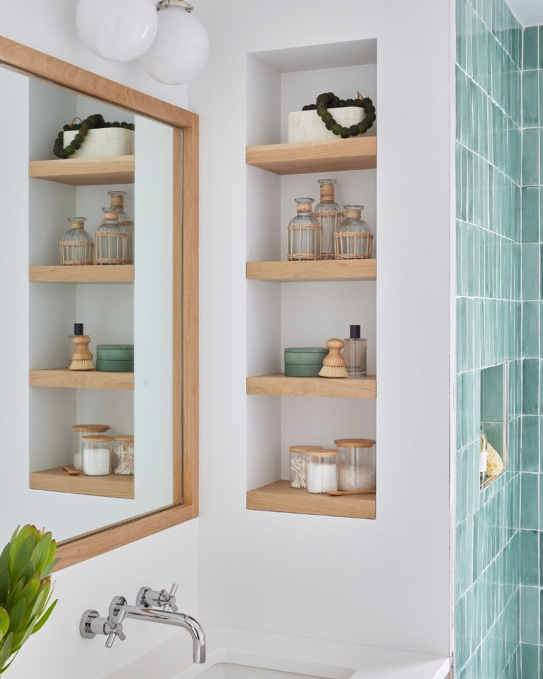 Four light natural wood floating shelves built into a white wall niche, holding clear glass bottles, green jars, and bath brushes.