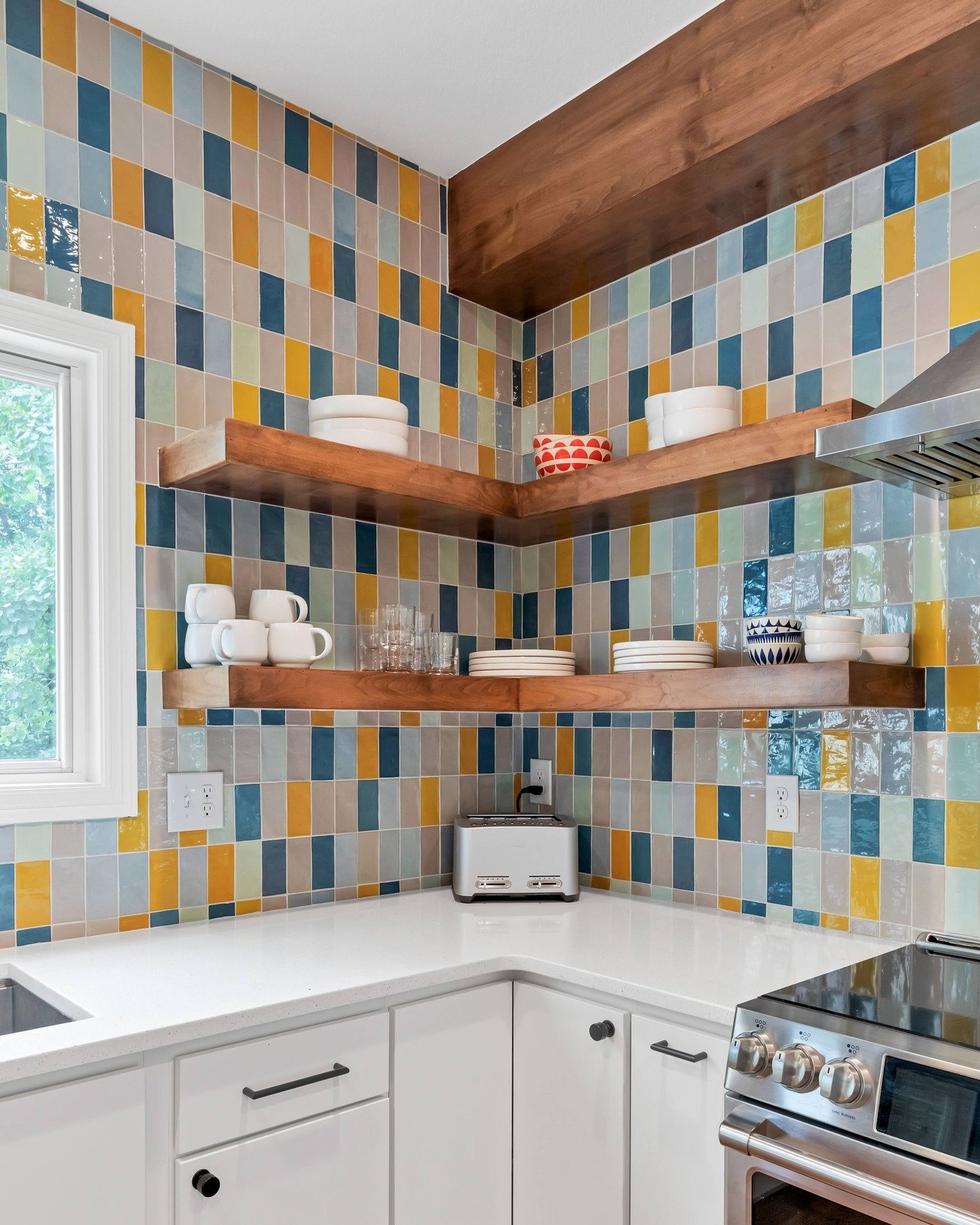 Kitchen corner backsplash with a random mosaic pattern of rectangular tiles in blue, yellow, gray, and white, contrasted with white cabinets and floating wooden shelves.