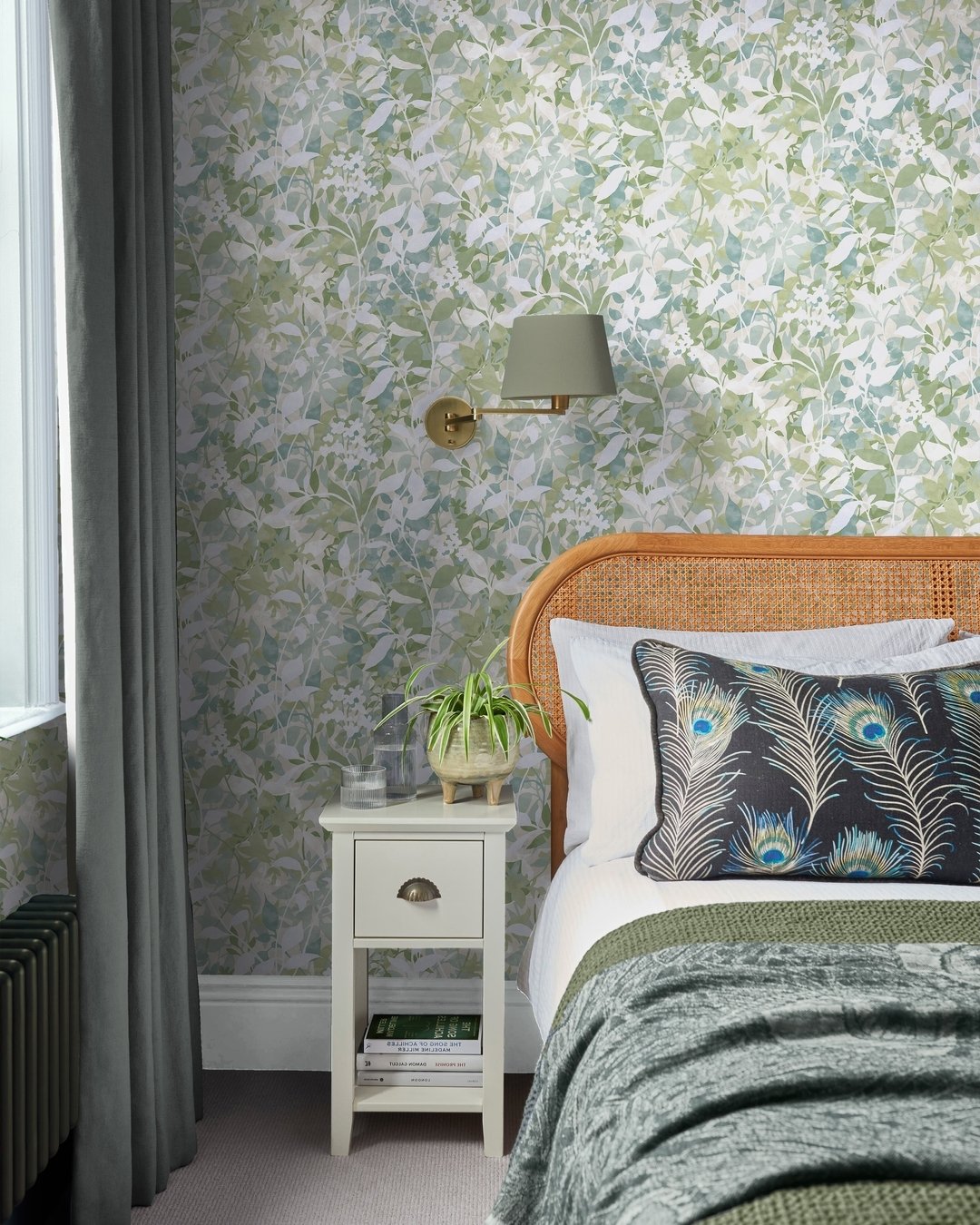 Bedroom with a striking sage green botanical print wallpaper, a cane headboard, and a peacock feather accent pillow.