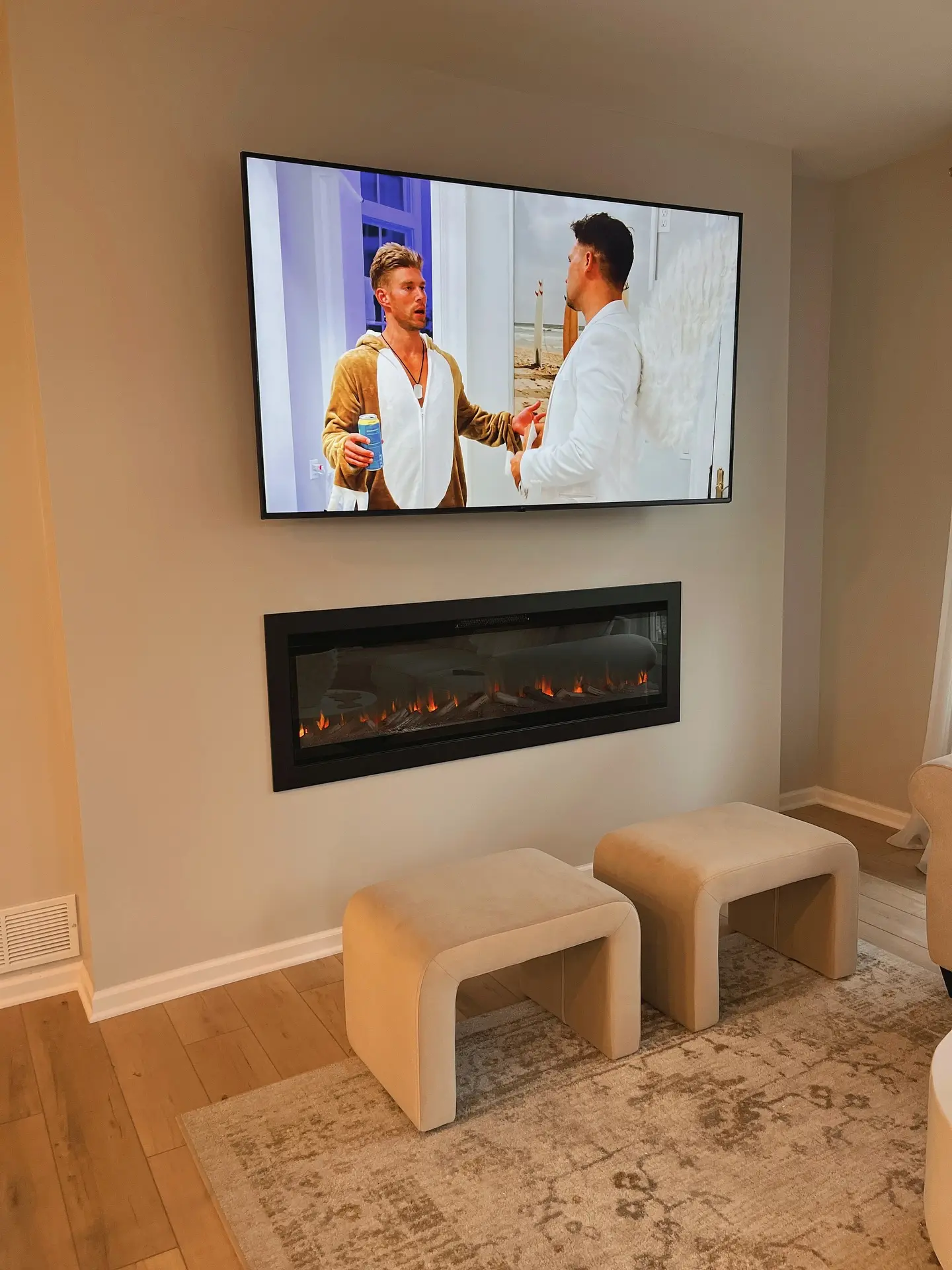 Minimalist living room with a large wall-mounted flatscreen TV directly above a flush, recessed linear electric fireplace on a light grey wall, with two beige upholstered ottomans below.
