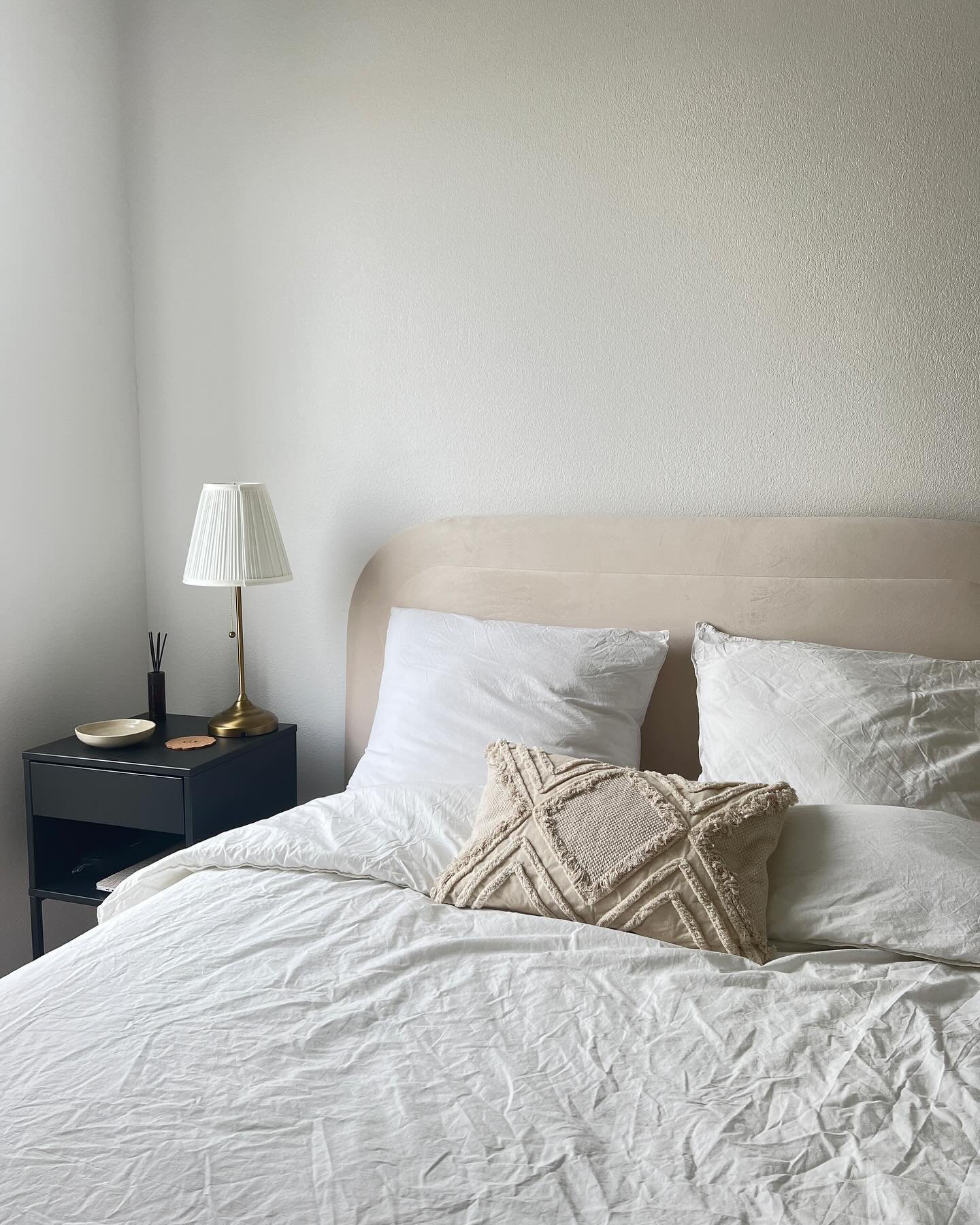 A contemporary minimalist bedroom corner featuring a low-profile bed with a rounded beige headboard, wrinkled white bedding, and a black metal nightstand with a gold-accented table lamp.