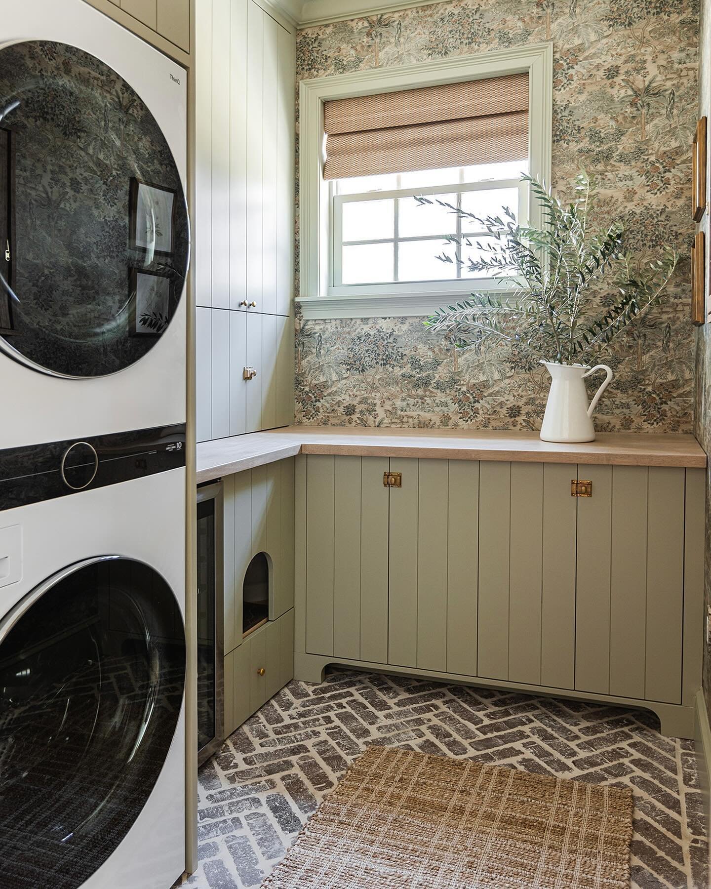 A small laundry room featuring a stacked washer and dryer, sage green beadboard-front cabinets, a busy woodland wallpaper, and a brick-look floor laid in a herringbone pattern. A custom cabinet includes a curved opening for a pet bed or litter box.