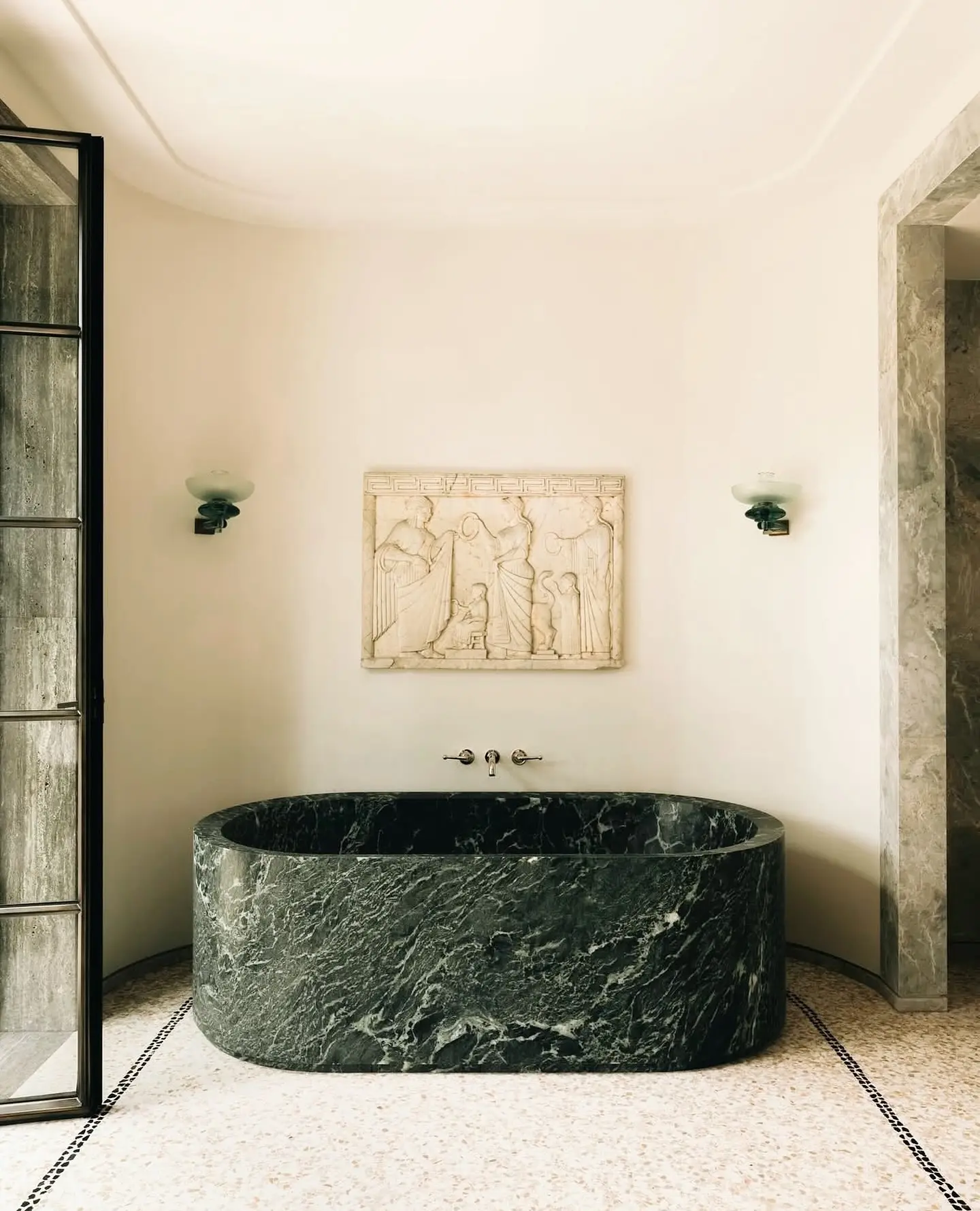 A luxurious bathroom with a large, oval dark green marble tub, featuring a light-colored classical bas-relief sculpture mounted on the wall above the faucet.