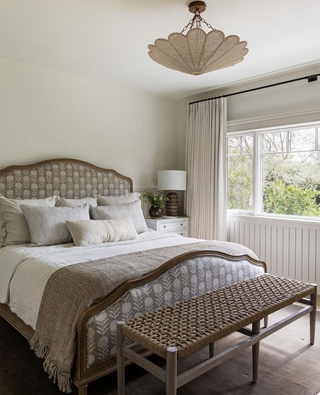 Neutral farmhouse bedroom with scalloped upholstered headboard, vertical wall paneling, and a woven bench at the foot of the bed.