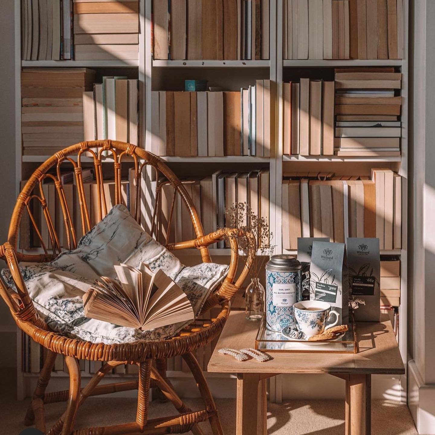 Bohemian reading nook with a natural woven rattan armchair, a light wood side table, and a wall of built-in bookshelves with sun shining through.