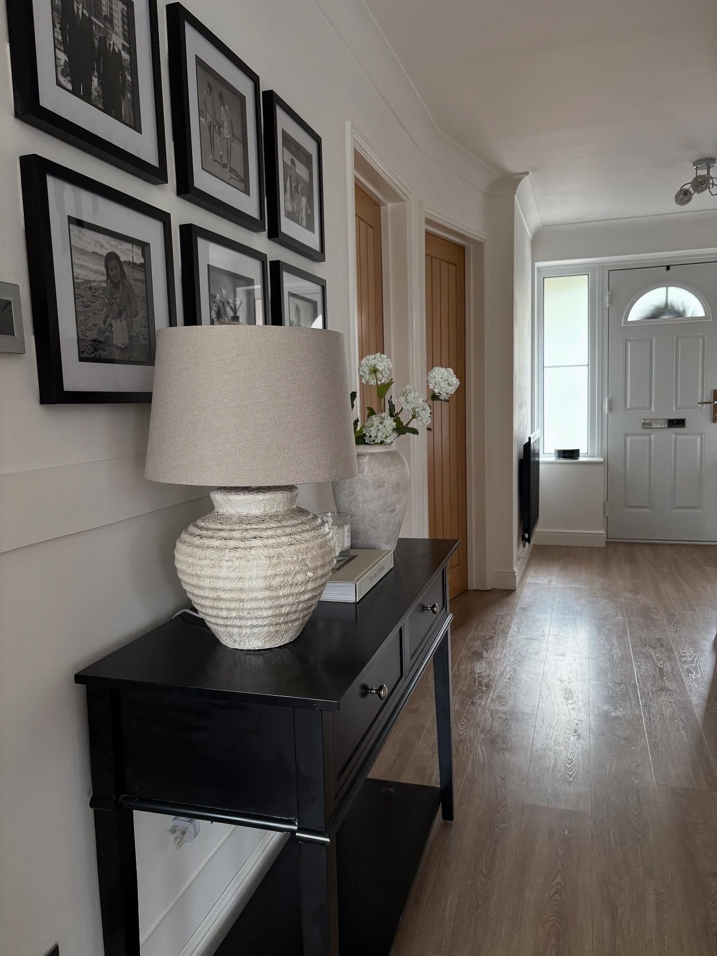 A black console table with drawers and a shelf, topped with a textured white lamp and white flowers, sitting below a grid of six framed black and white photos.