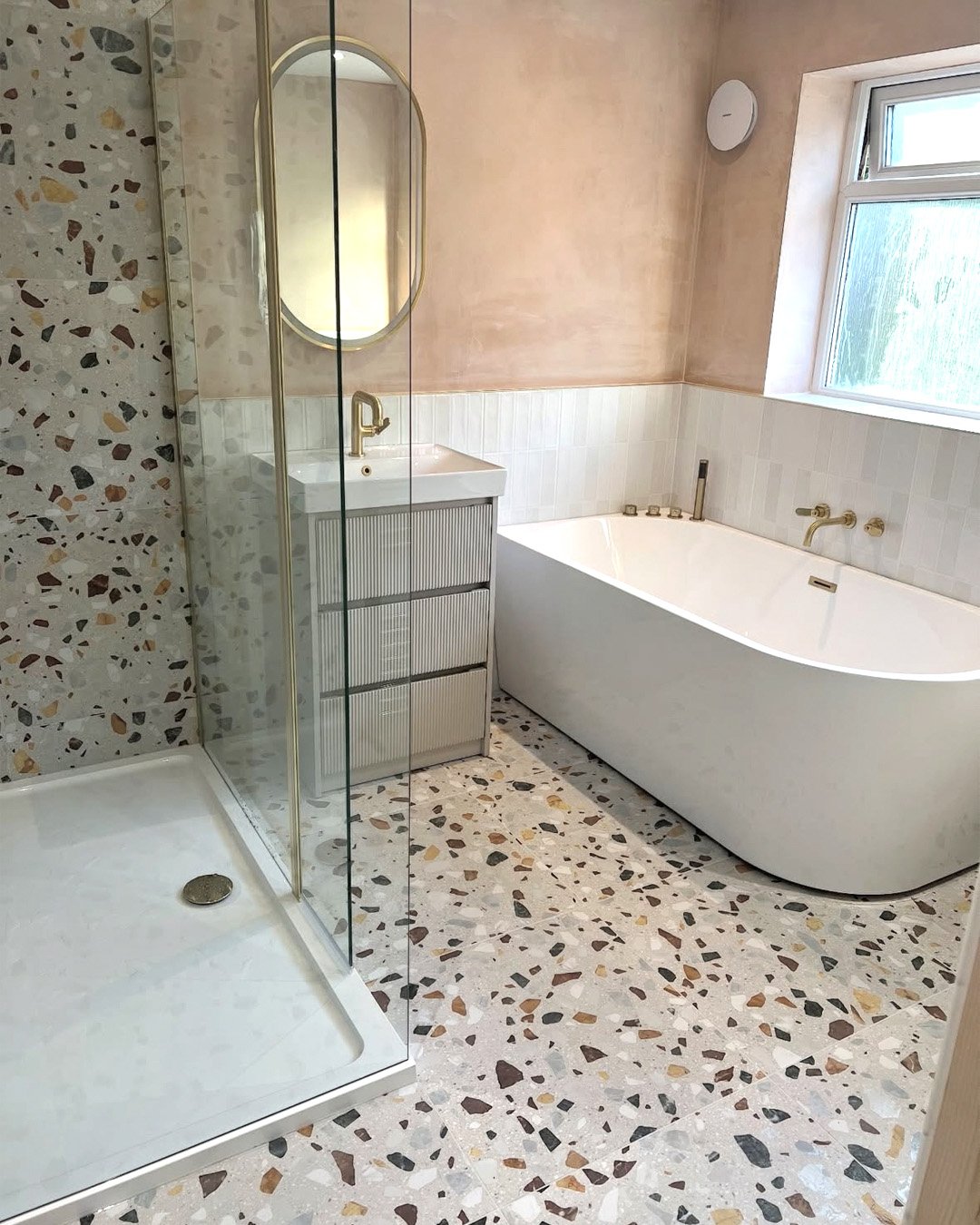 A small, contemporary bathroom with a white freestanding tub and a floor and shower wall covered in light gray terrazzo tile with large, colorful stone fragments.