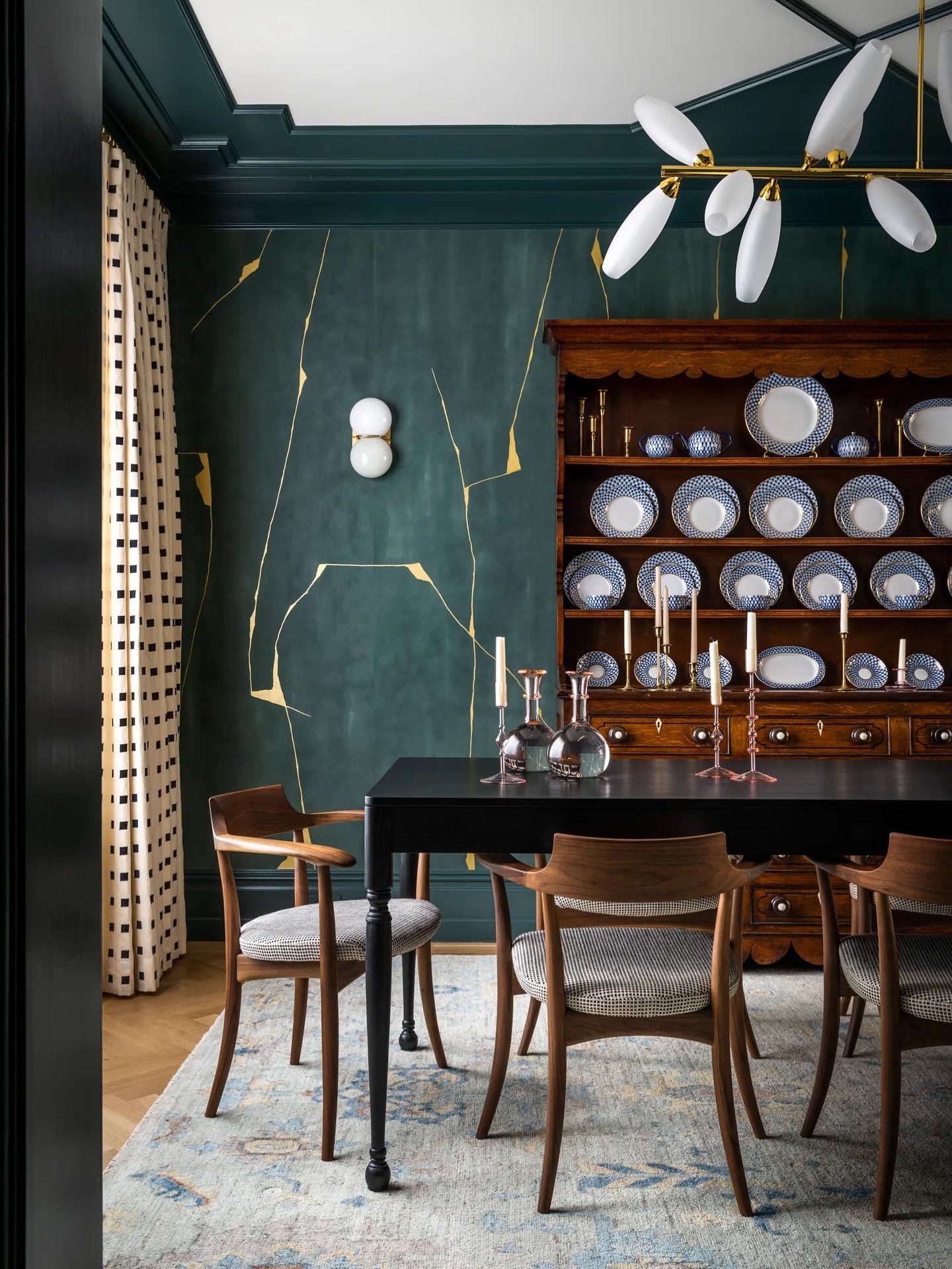 A dramatic dining room featuring a dark teal wall with gold leaf kintsugi-style accents, an antique wooden china cabinet, a black table, and mid-century wooden chairs.