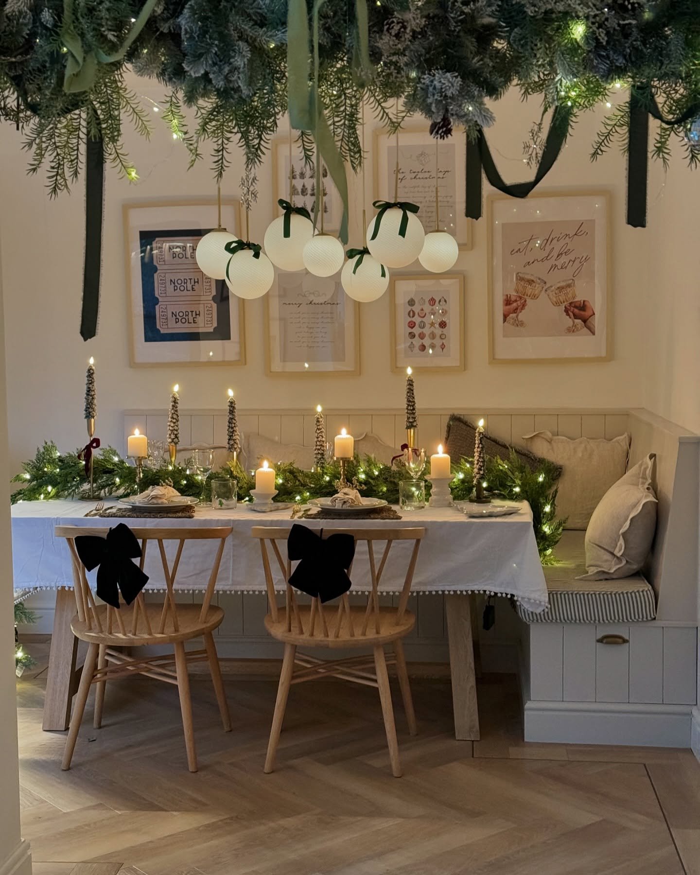 A cozy breakfast nook table set with a white tablecloth, a lush evergreen garland, thin gold taper candles, and light wood chairs decorated with large black velvet bows. Green ribbons hang from an overhead floral display.