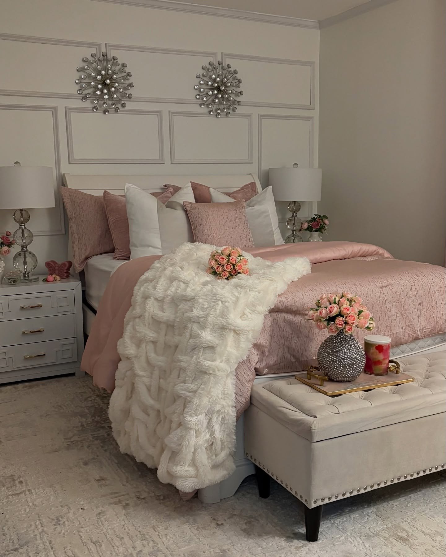Symmetrical pink and white bedroom with paneled accent wall, mirrored nightstands, and two large crystal starburst wall sconces.