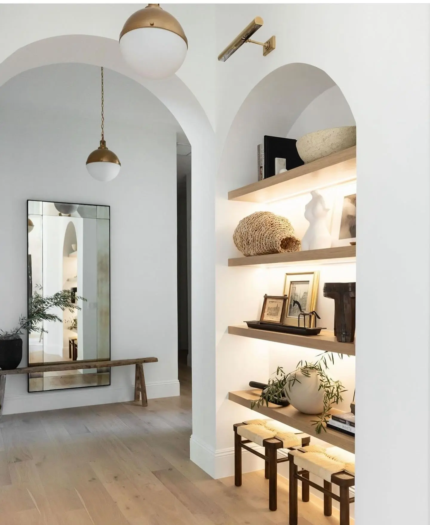 Modern white hallway with light wood flooring, arched display niches with backlit floating shelves, brass and opal glass pendants, and a slim brass picture light sconce.