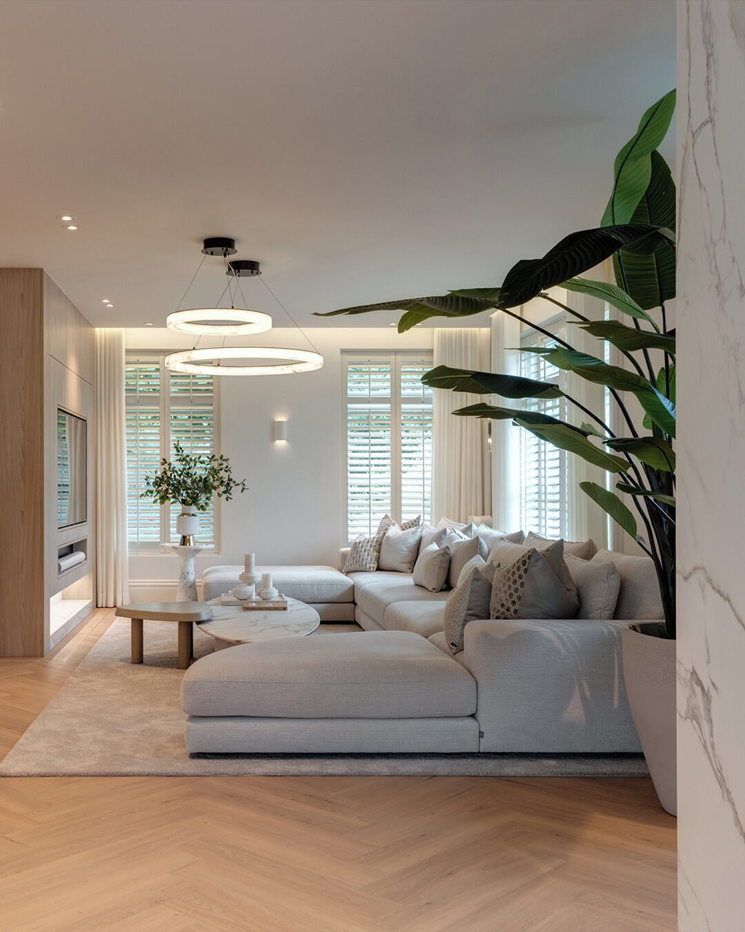 A luxurious, light beige living room featuring a large U-shaped sectional sofa, white plantation shutters, chevron wood flooring, and modern circular pendant lights.