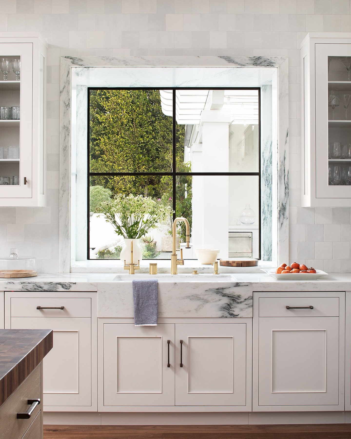 White transitional kitchen with cream cabinets. A large black-framed picture window is recessed and trimmed entirely in matching gray and white marble, centered above a marble farmhouse sink.