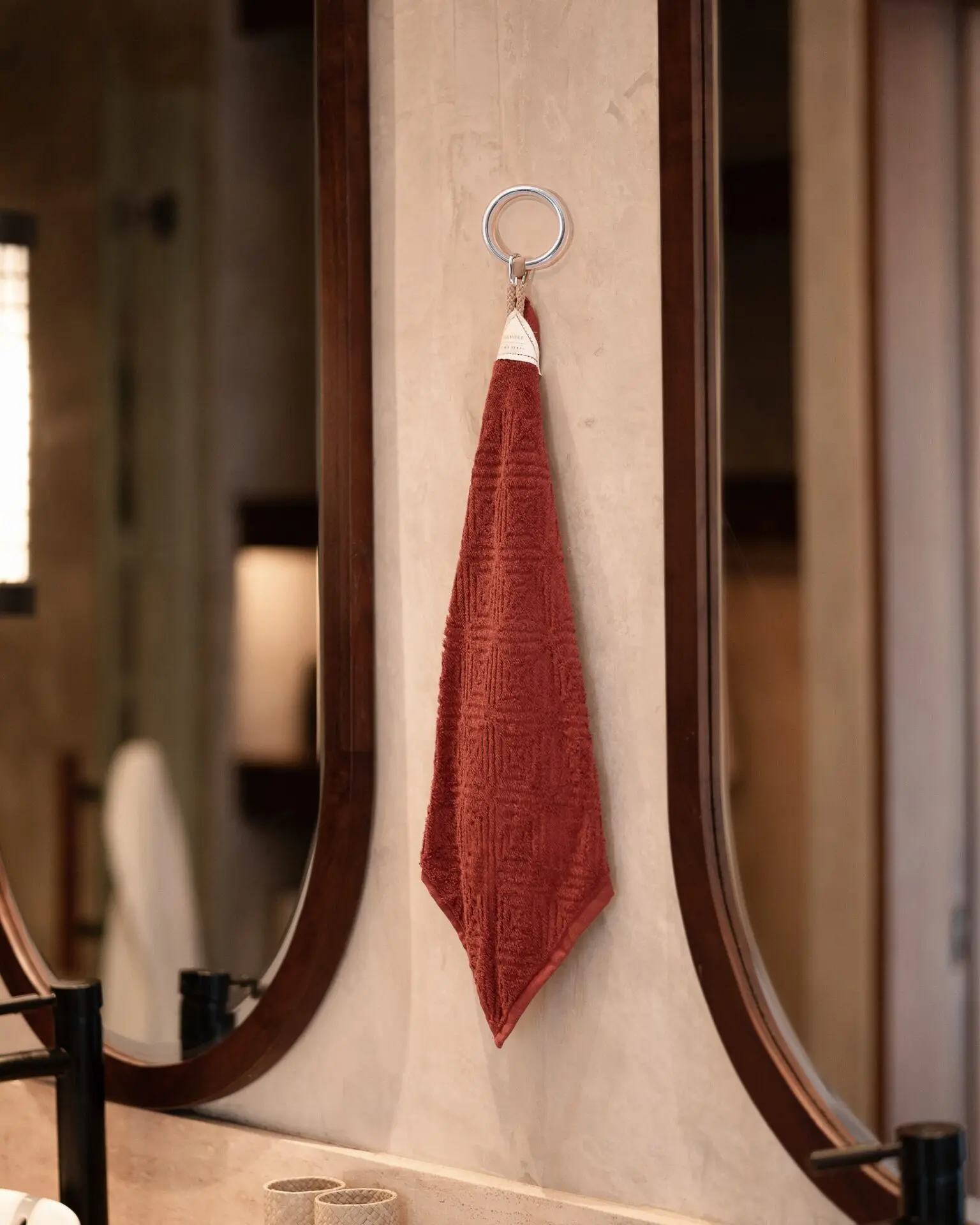 A small silver metal keyring or clip holding a terracotta red hand towel by its loop on a beige wall between two dark wood mirrors.