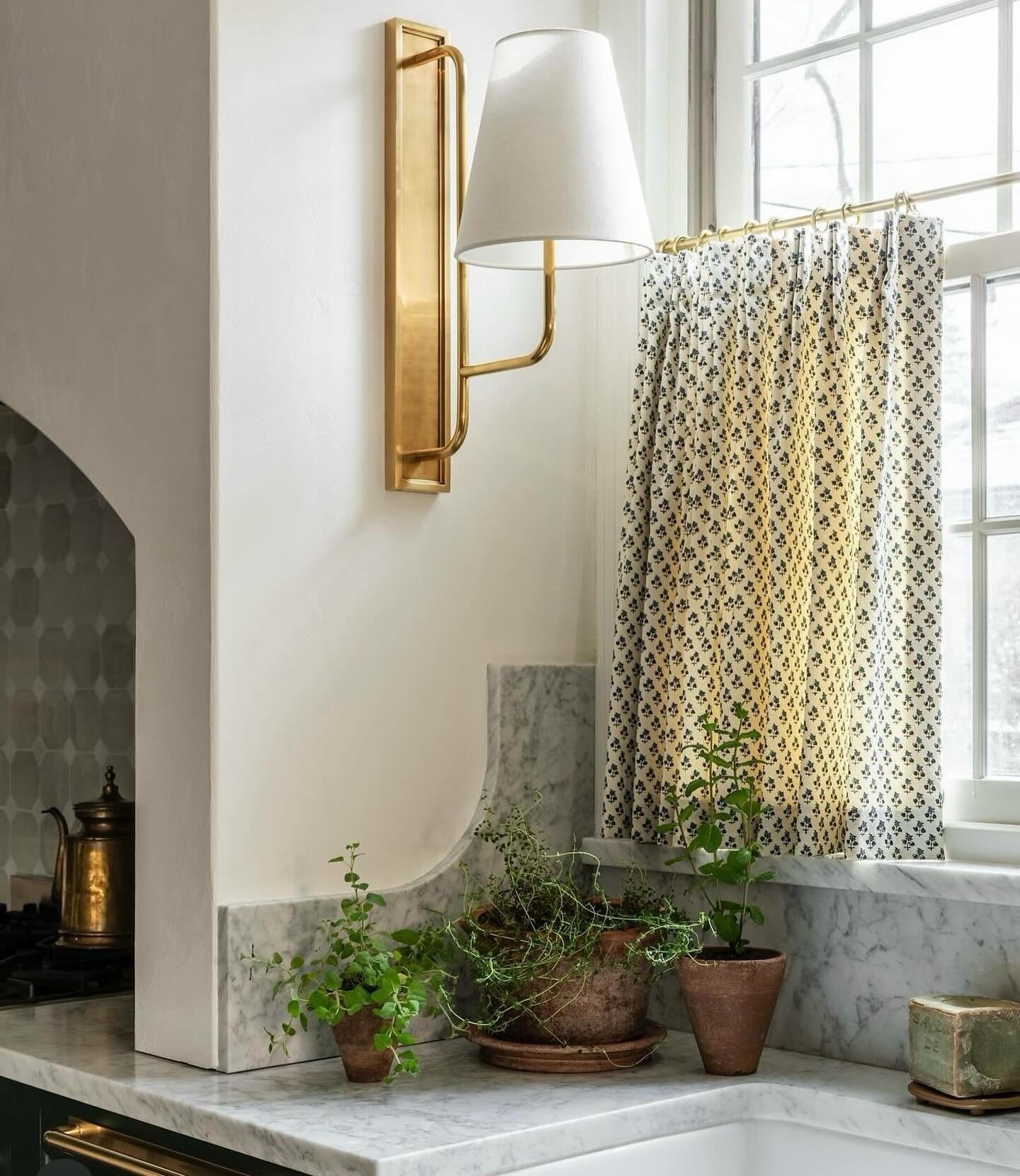 Kitchen sink area with a marble countertop, featuring a small cafe curtain in a black and white repeating small print pattern hung on a brass rod.
