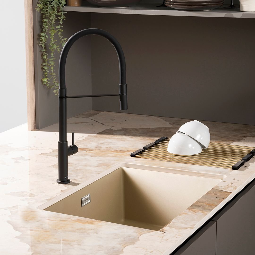 A close-up of a square, single-bowl beige or tan granite composite undermount sink set in a brown and beige marbled quartz countertop, featuring a matte black flexible spout faucet and a black and gold roll-up drying rack.