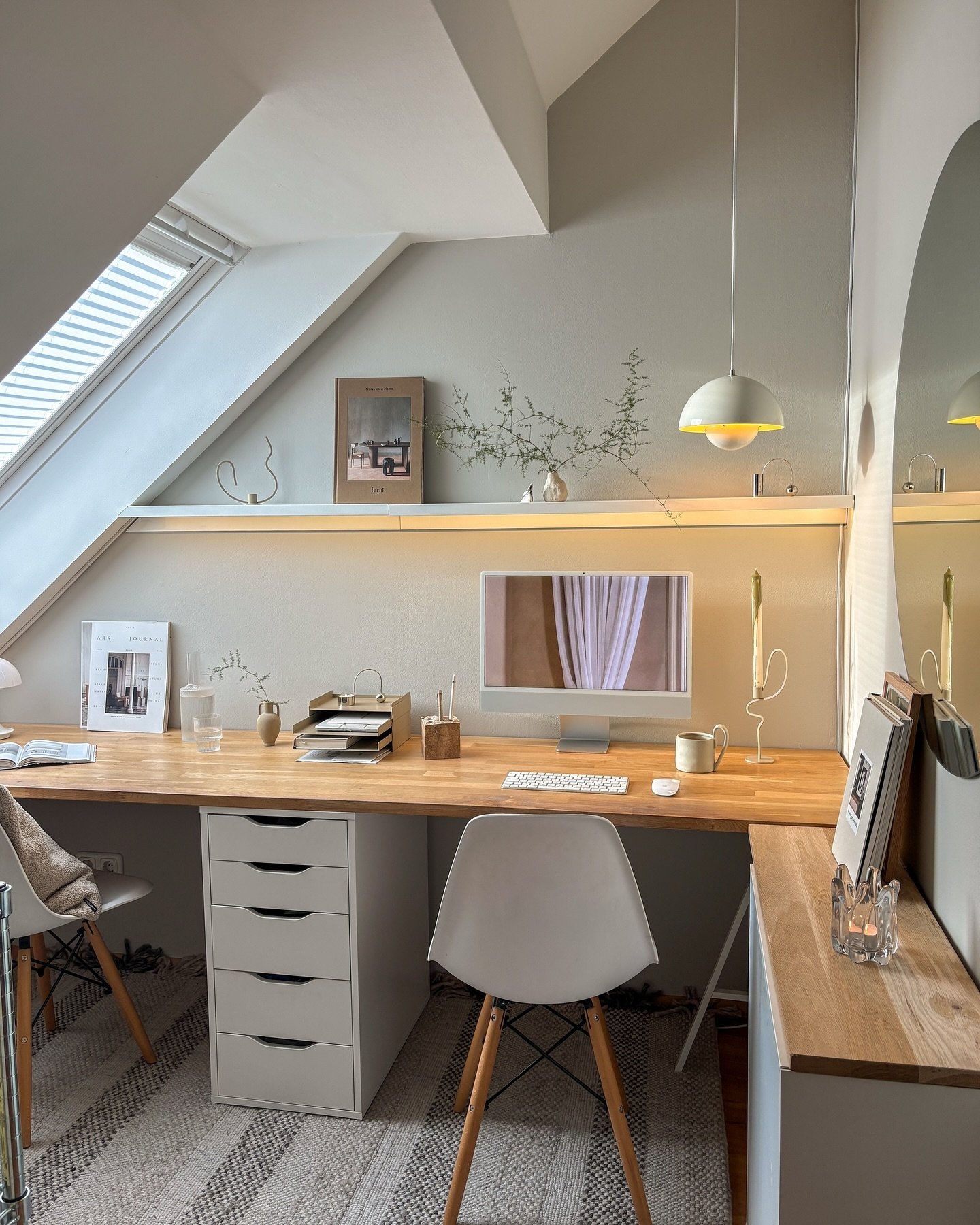 Cozy attic home office with sloped ceilings, a long light wood desk, white drawer units, two white chairs, and a small white pendant light.
