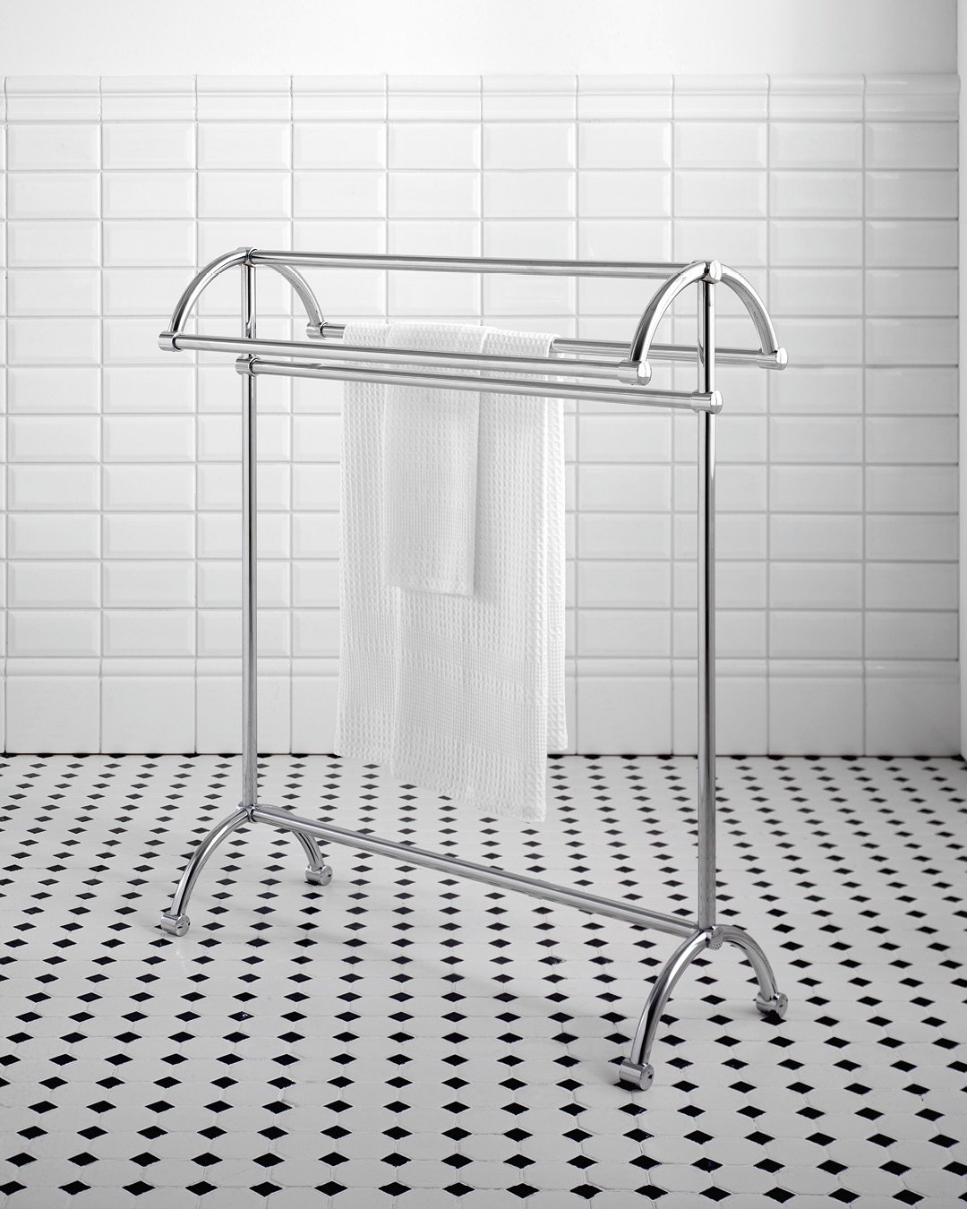 Freestanding, polished chrome towel rack with an arched top and two hanging bars, holding a white towel in a bathroom with white subway tiles and black and white floor tiles.