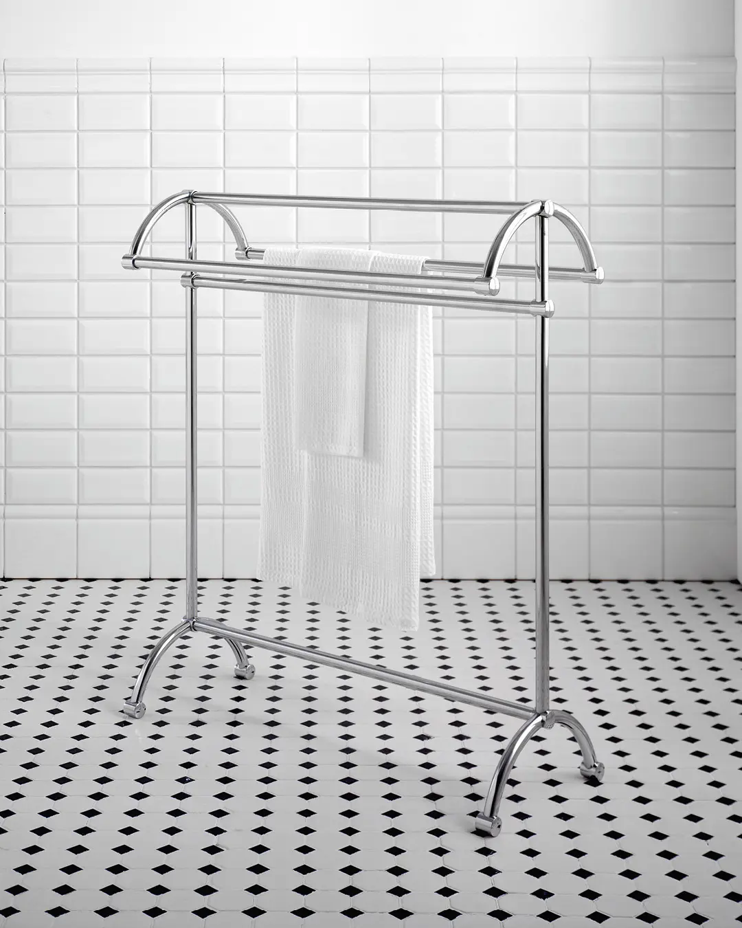 Freestanding, polished chrome towel rack with an arched top and two hanging bars, holding a white towel in a bathroom with white subway tiles and black and white floor tiles.