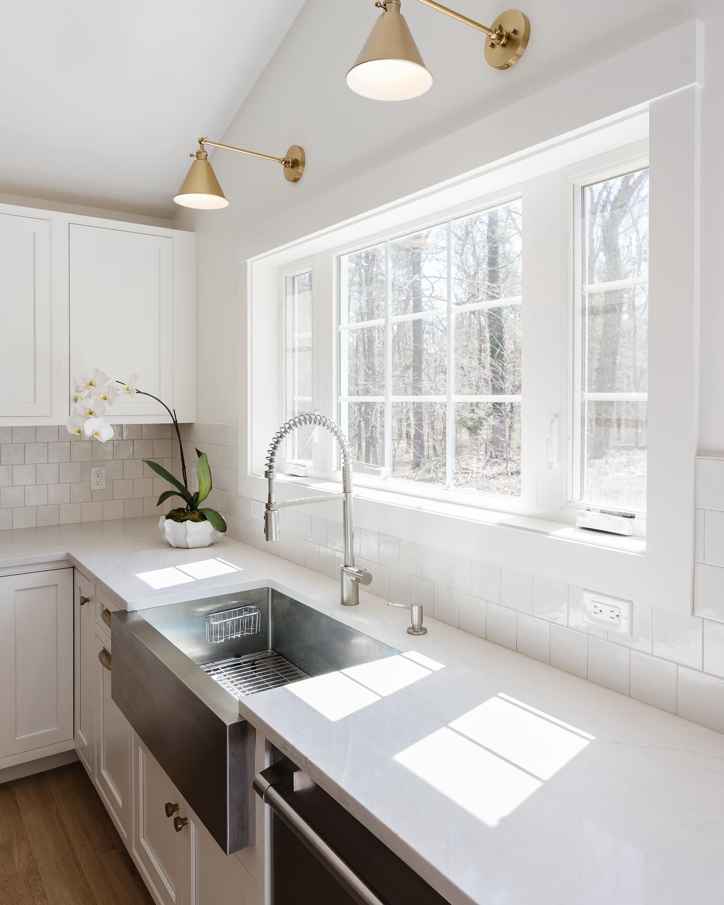 Modern white kitchen with subway tile backsplash and white quartz counters. A large stainless steel apron-front sink is positioned below a bright triple window set with white frames.