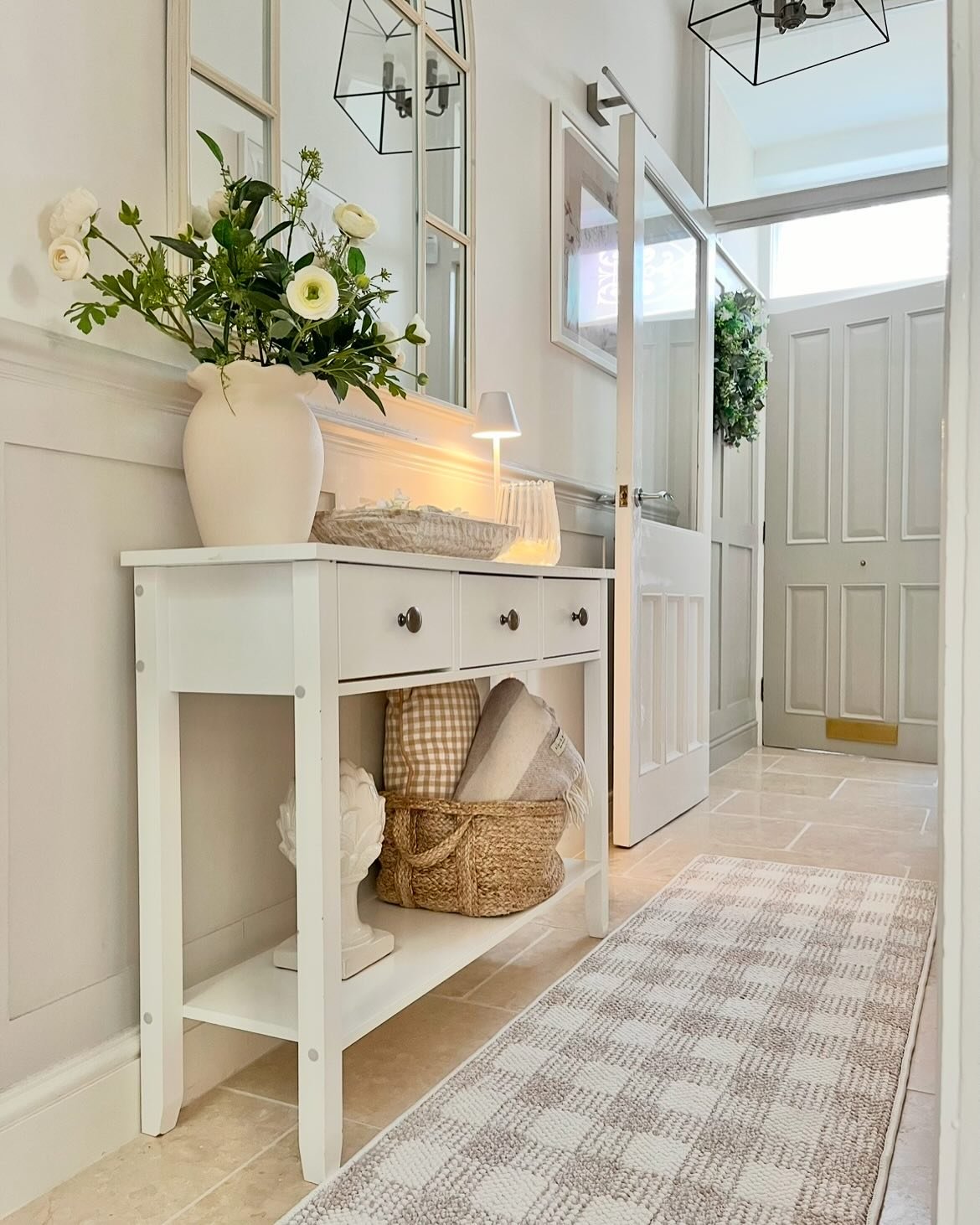 A white console table with three drawers in a light grey hallway, topped with white flowers and a small lamp, with a large mirror leaning above it.