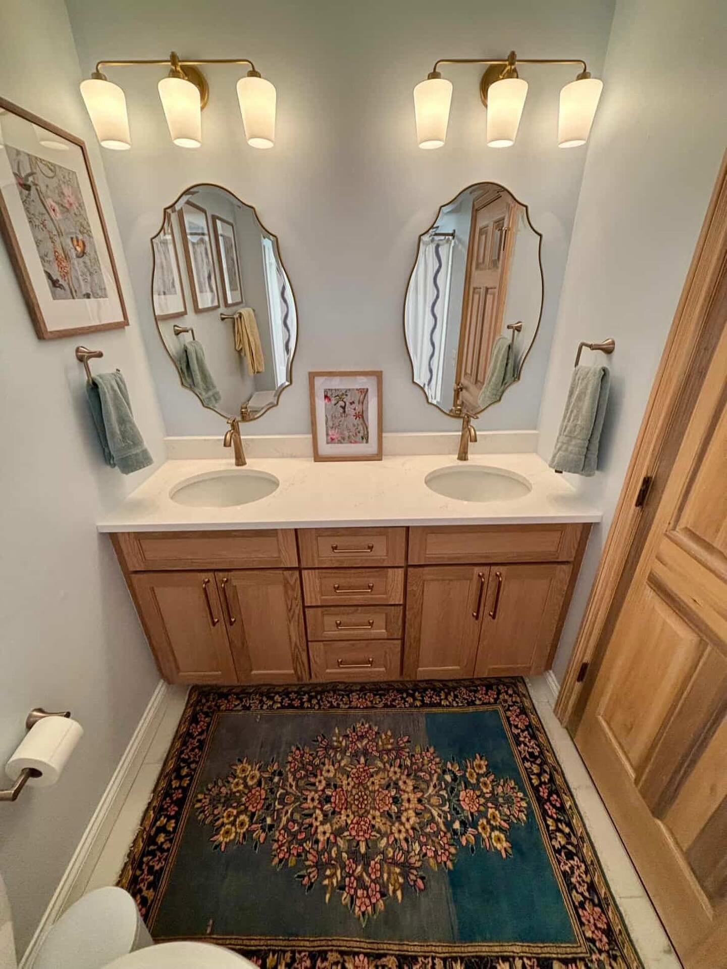 A dual vanity area featuring a light wood cabinet, brass fixtures, scalloped oval mirrors, and a rectangular rug with a prominent floral medallion pattern on a deep teal and blue background.