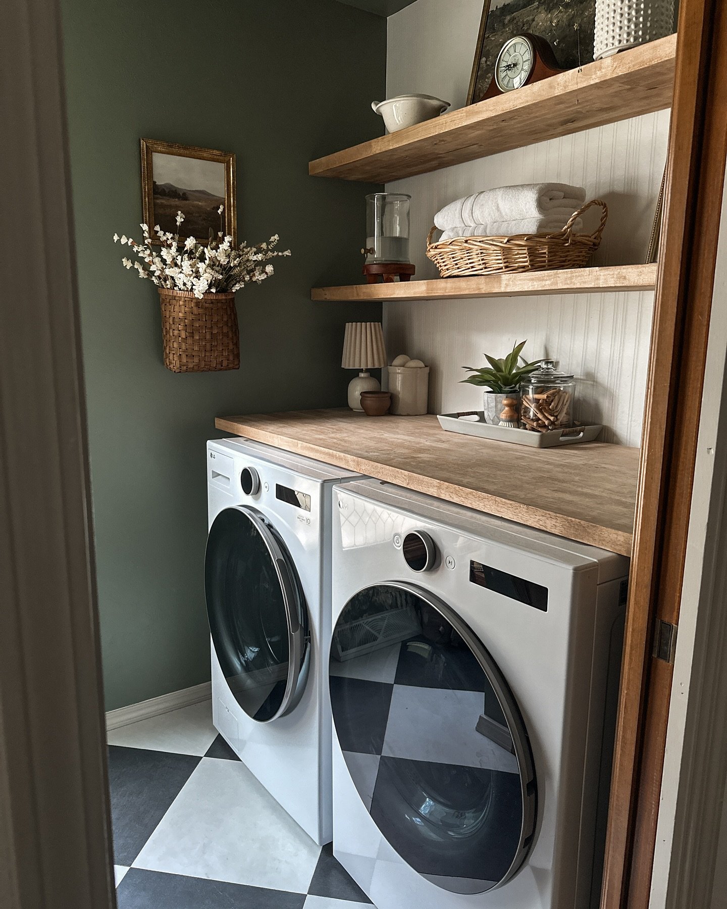 A tiny laundry closet featuring walls painted a deep olive green, a light wood countertop over the washer and dryer, open shelving on a beadboard back panel, and a black and white checkerboard floor.