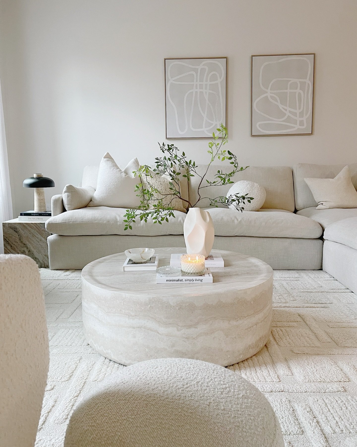 A neutral living room featuring a massive round travertine coffee table, a low-slung beige sofa, and a pair of minimalist abstract line art prints.