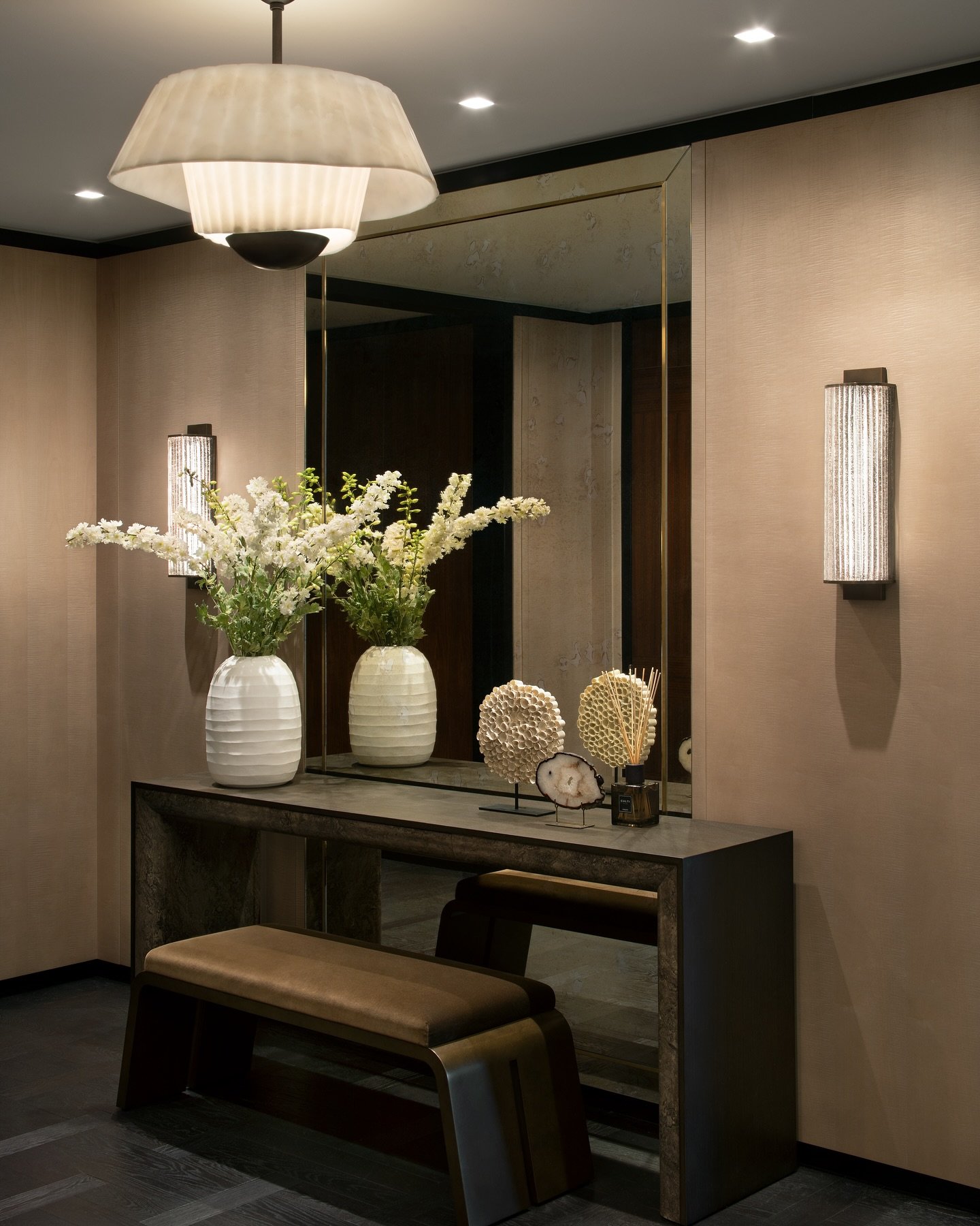 Luxurious, beige-toned entry hall with textured walls, a large bronze-framed mirror, two vertical ribbed glass wall sconces, and a tiered fabric ceiling light.