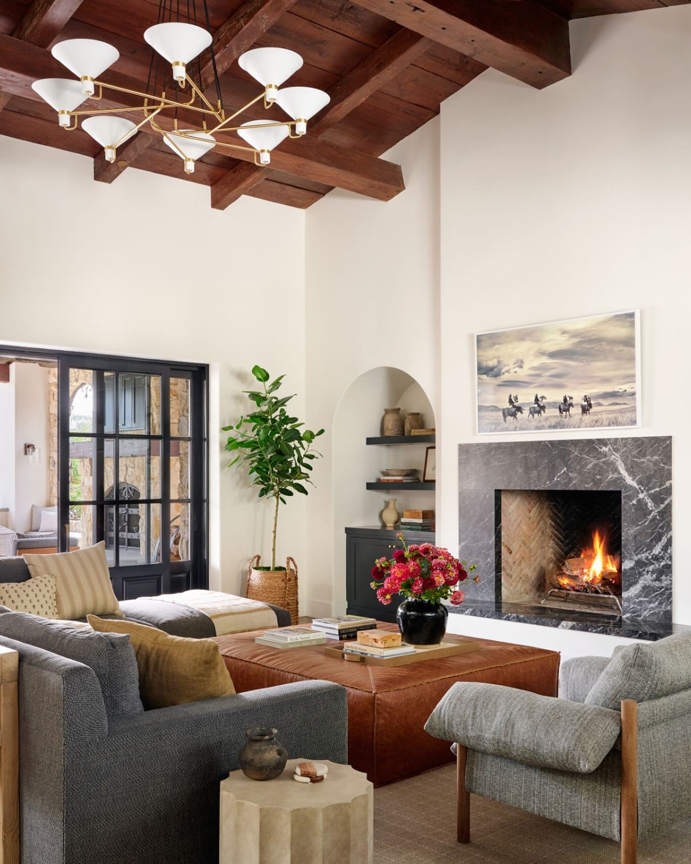 A cozy living room featuring dark wood ceiling beams, a striking black marble fireplace, a large square leather ottoman, and a gray sofa with a modern brass chandelier.
