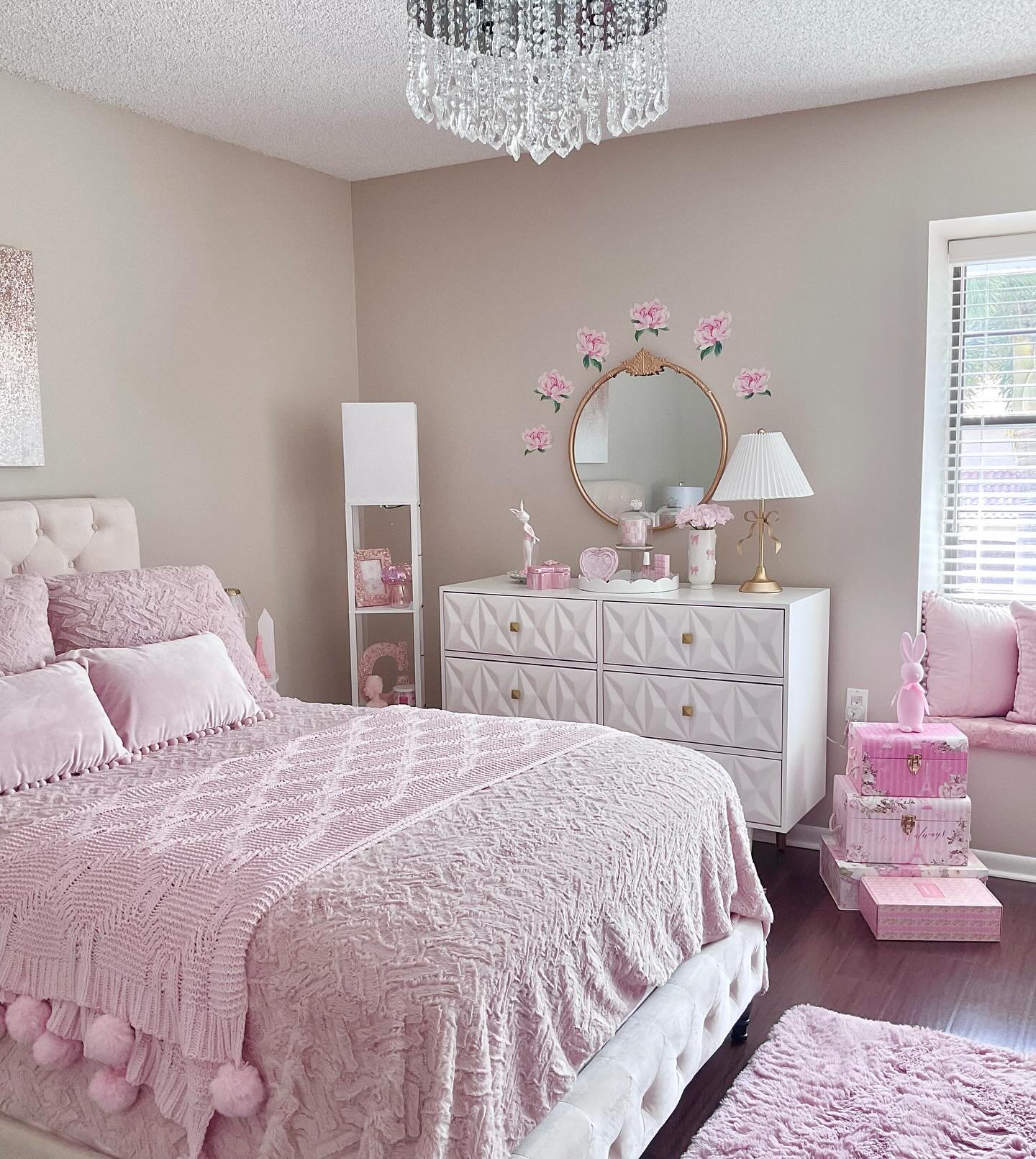 Bedroom with taupe walls, a white dresser with diamond-patterned drawers, a crystal chandelier, and a bed with a chunky knit pom-pom blanket.