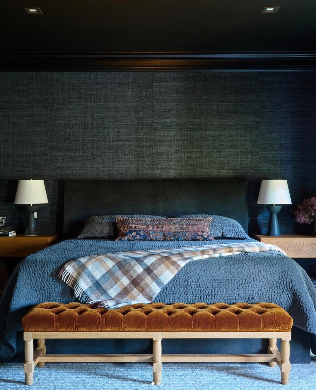 A dramatic, dark bedroom with a navy blue bedspread and a long bench at the foot of the bed, upholstered in rich, burnt-orange tufted velvet with light wood legs.