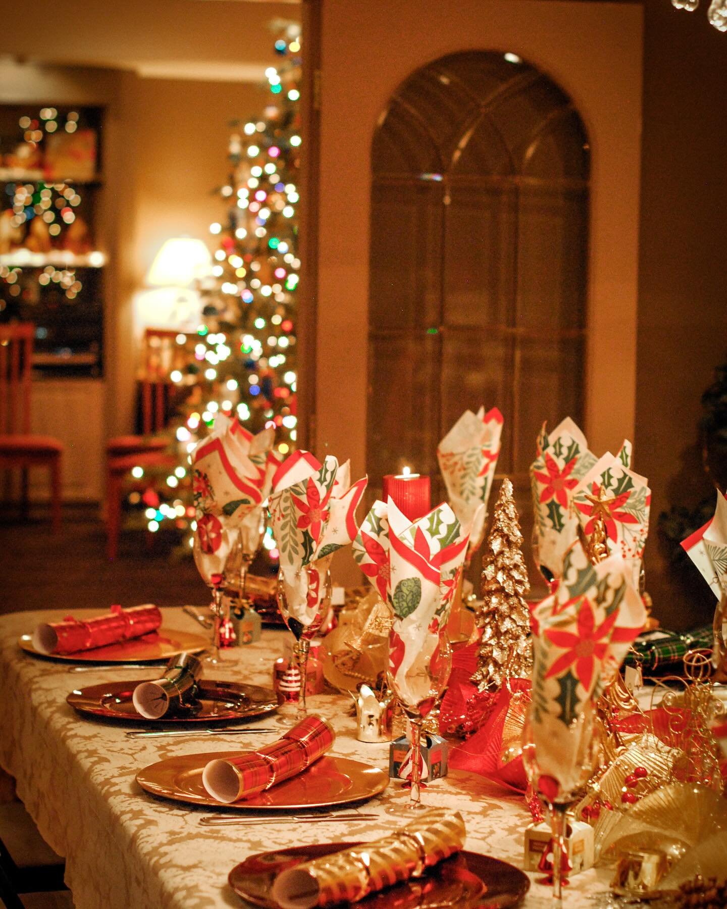A festive dining table with a cream damask tablecloth, set with shiny copper or gold charger plates, red and green patterned napkins folded upright, red and gold Christmas crackers, and a centerpiece of gold glitter trees.